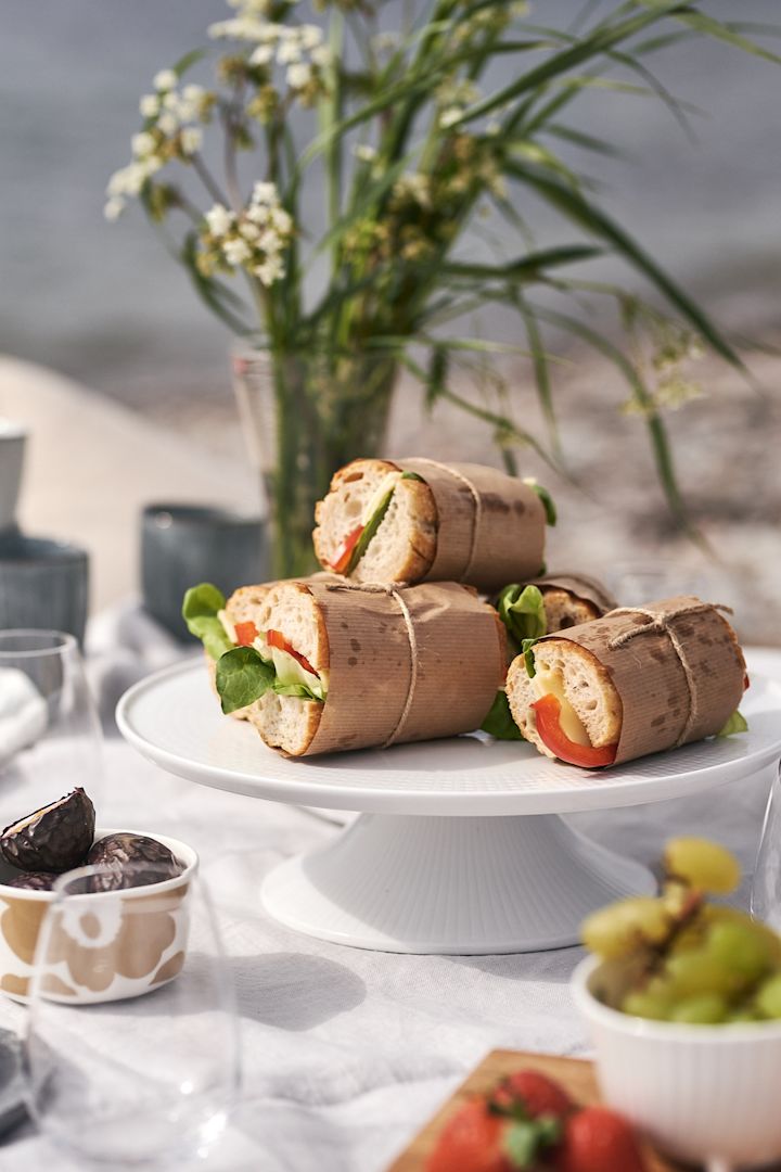 Piknik på stranden med smørbrød pakket inn i vokset papir på et hvitt kakefat.