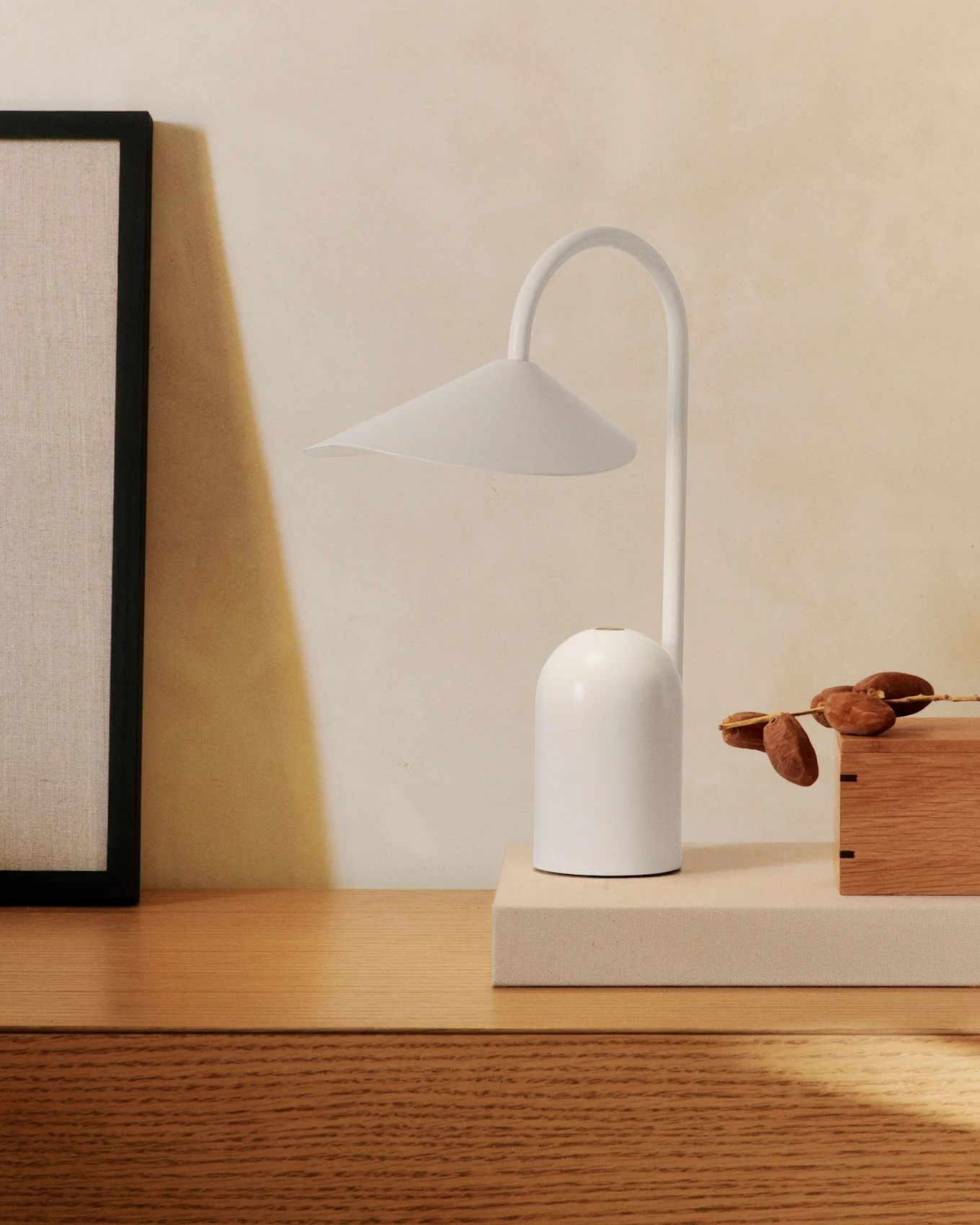 En minimalistisk hvit bordlampe med buet arm på en treskjenk, ved siden av et innrammet bilde og en trekasse med dadler.