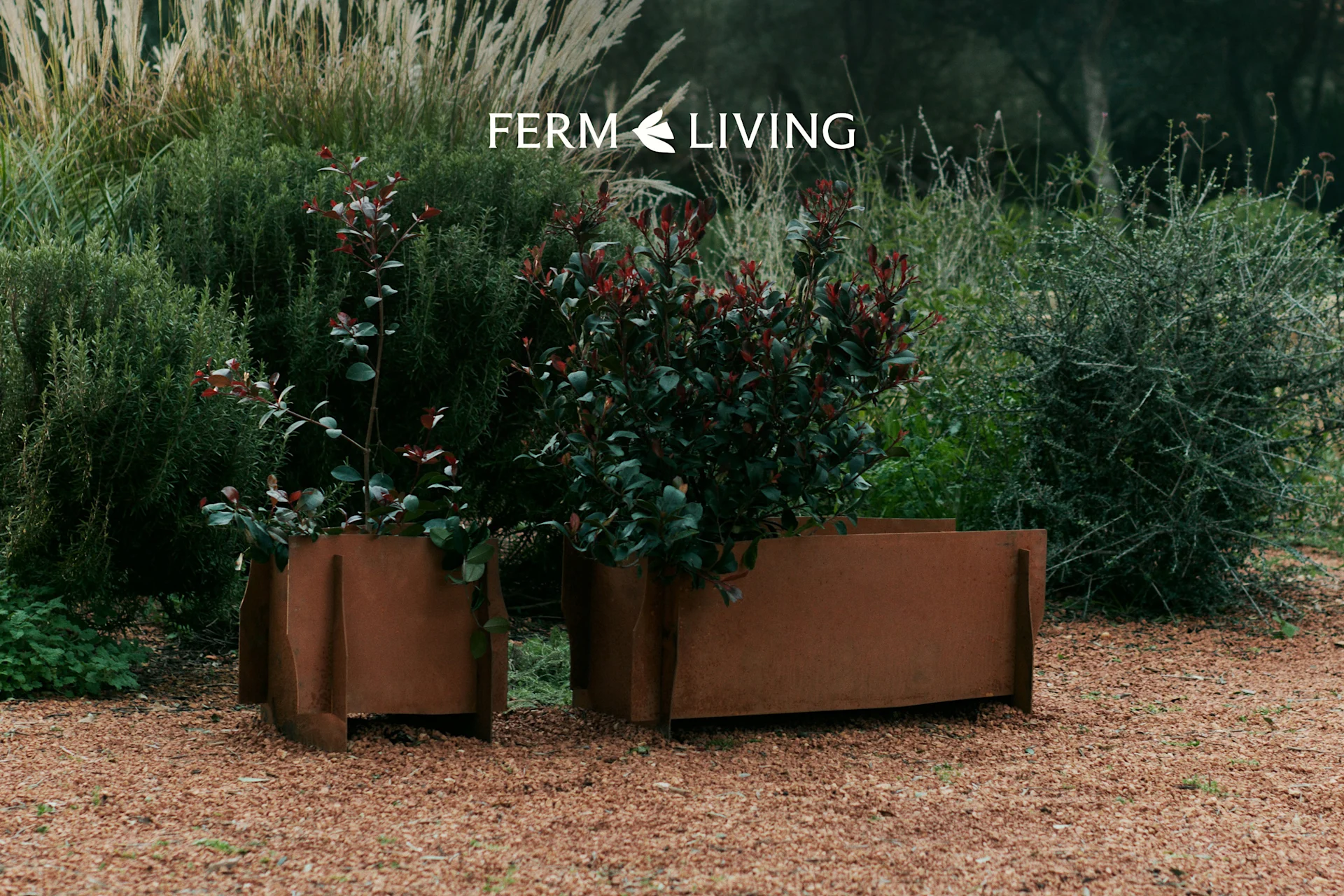 To værbitte metallplantekasser med grønne og røde blader på grus i en hage. Ferm Living-logo.