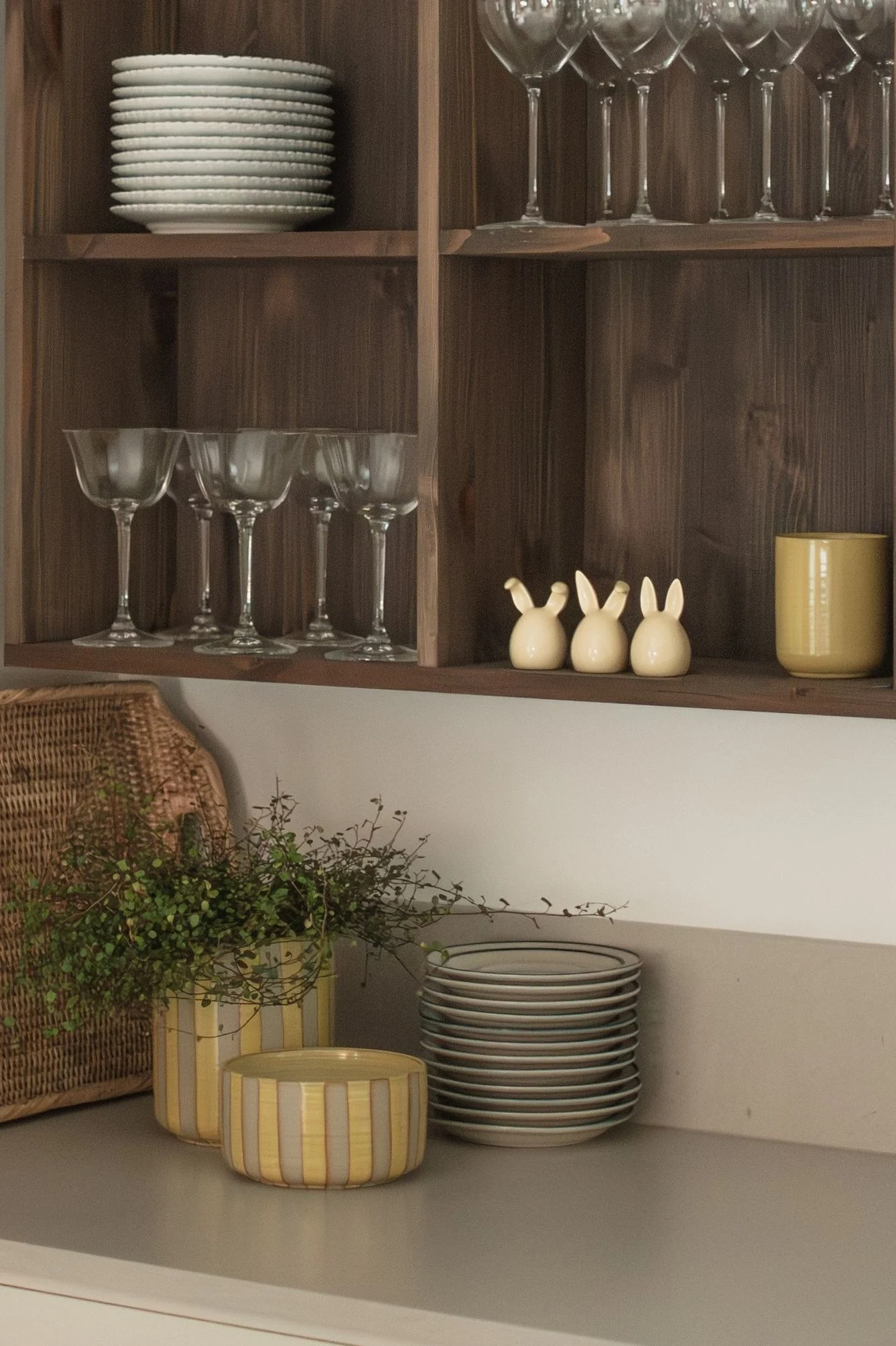 Trehyller med stablede hvite tallerkener, forskjellige glass, tre keramiske kaniner og et gult krus. På benken står en flettet kurv, planter i stripete potter og flere tallerkener.