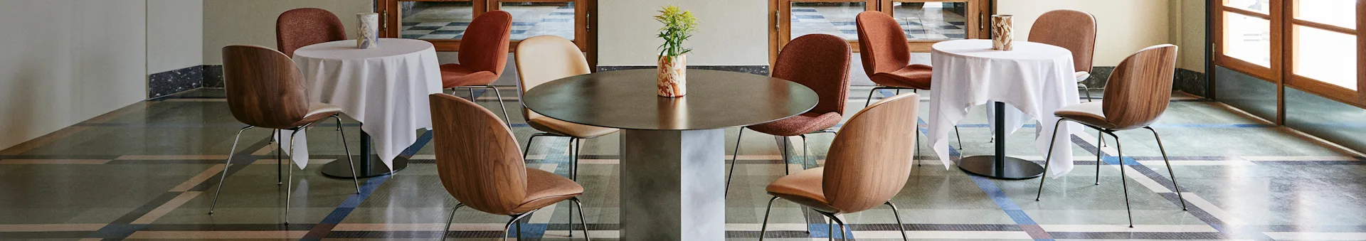 Et stilfullt kaféinteriør med et stort sentralt bord, to mindre bord med hvite duker og diverse stoler på et mønstret gulv.