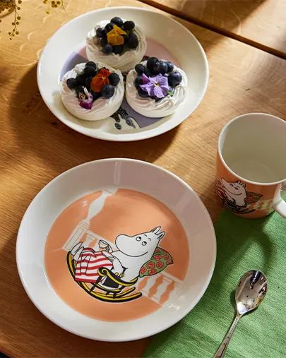 Moomintroll-tallerken, krus og skje ved siden av en tallerken med marengs med blåbær og blomster på et trebord.