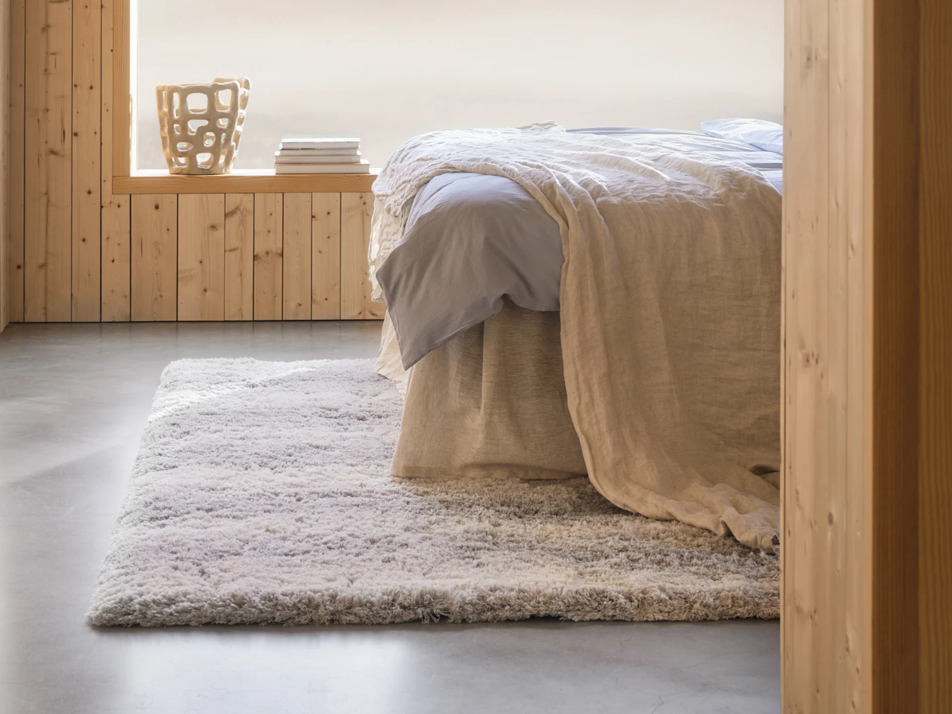 Koselig soverom med seng, lyseblå laken, beige teppe, lurvete gulvteppe, trepanelvegger og en dekorativ vase.