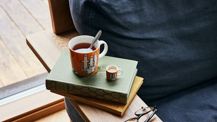 Den oransje Mummikoppen med Hatifnatten og te står ved siden av en sofa, oppå en stabel med bøker.
