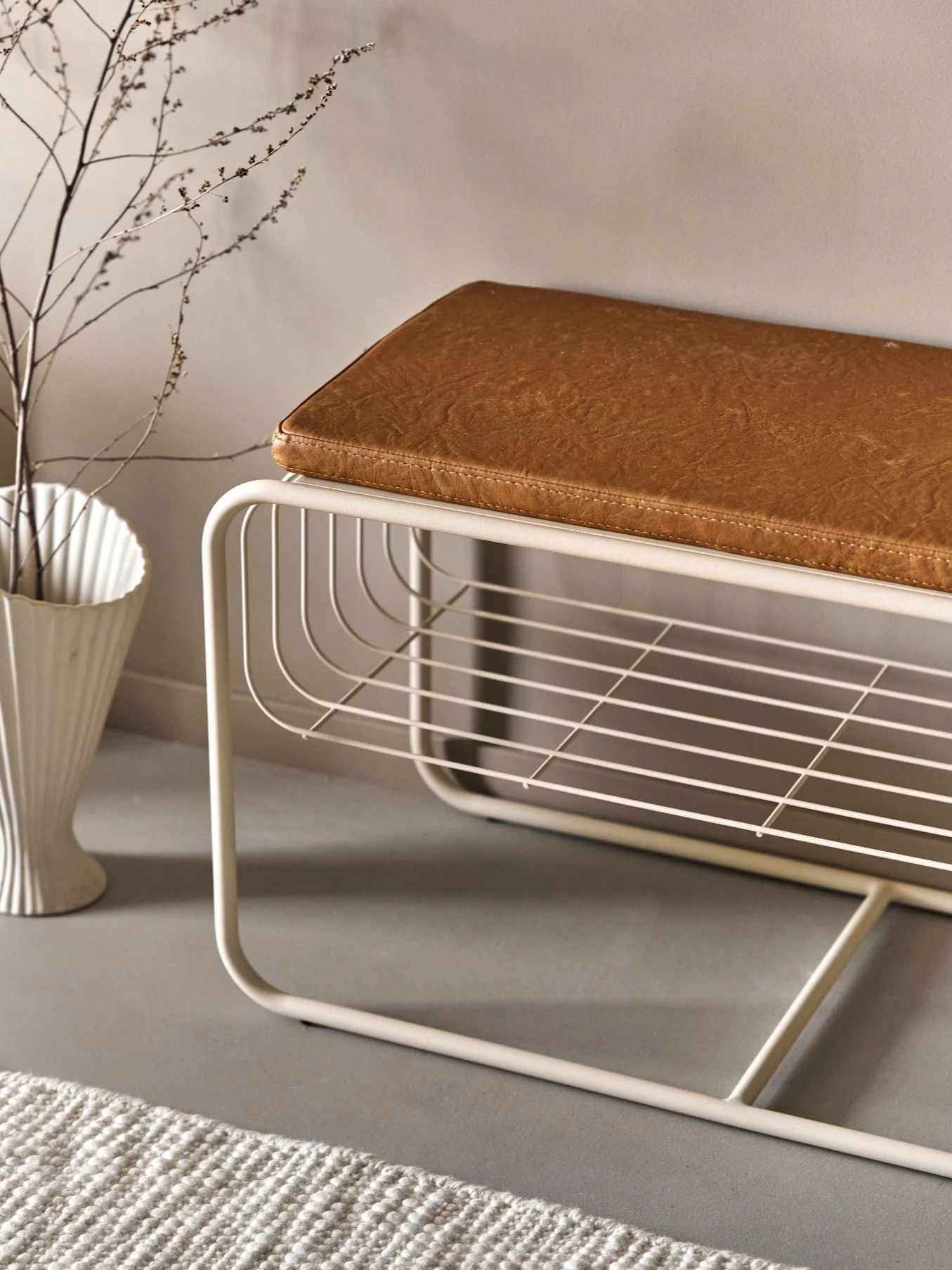 Moderne beige metallbenk med brun kunstskinnpute og åpen spilehylle. En hvit vase med grener og et teppe er i nærheten.