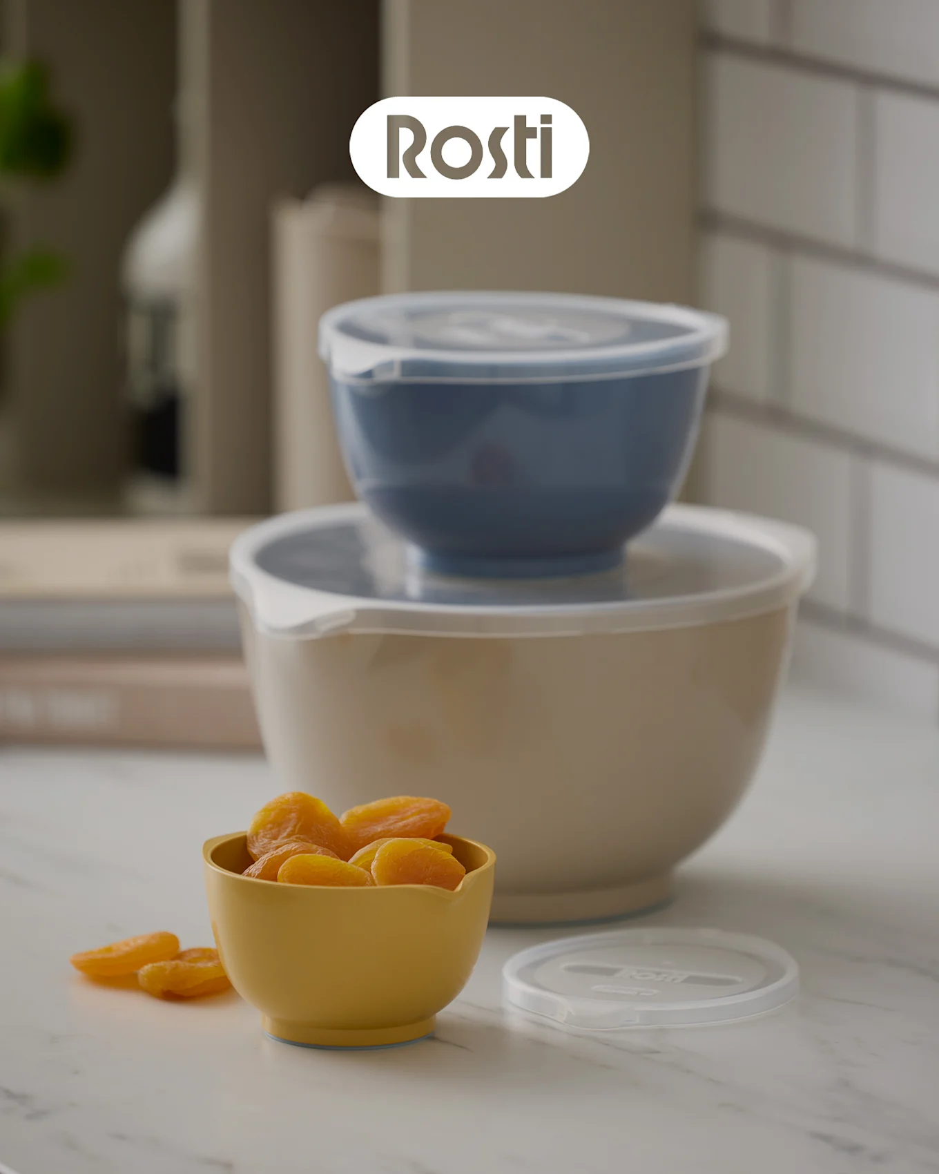 Tre Rosti-skåler på en benk: en beige skål med en blå stablet oppå, begge med lokk, og en liten gul skål fylt med tørkede aprikoser.