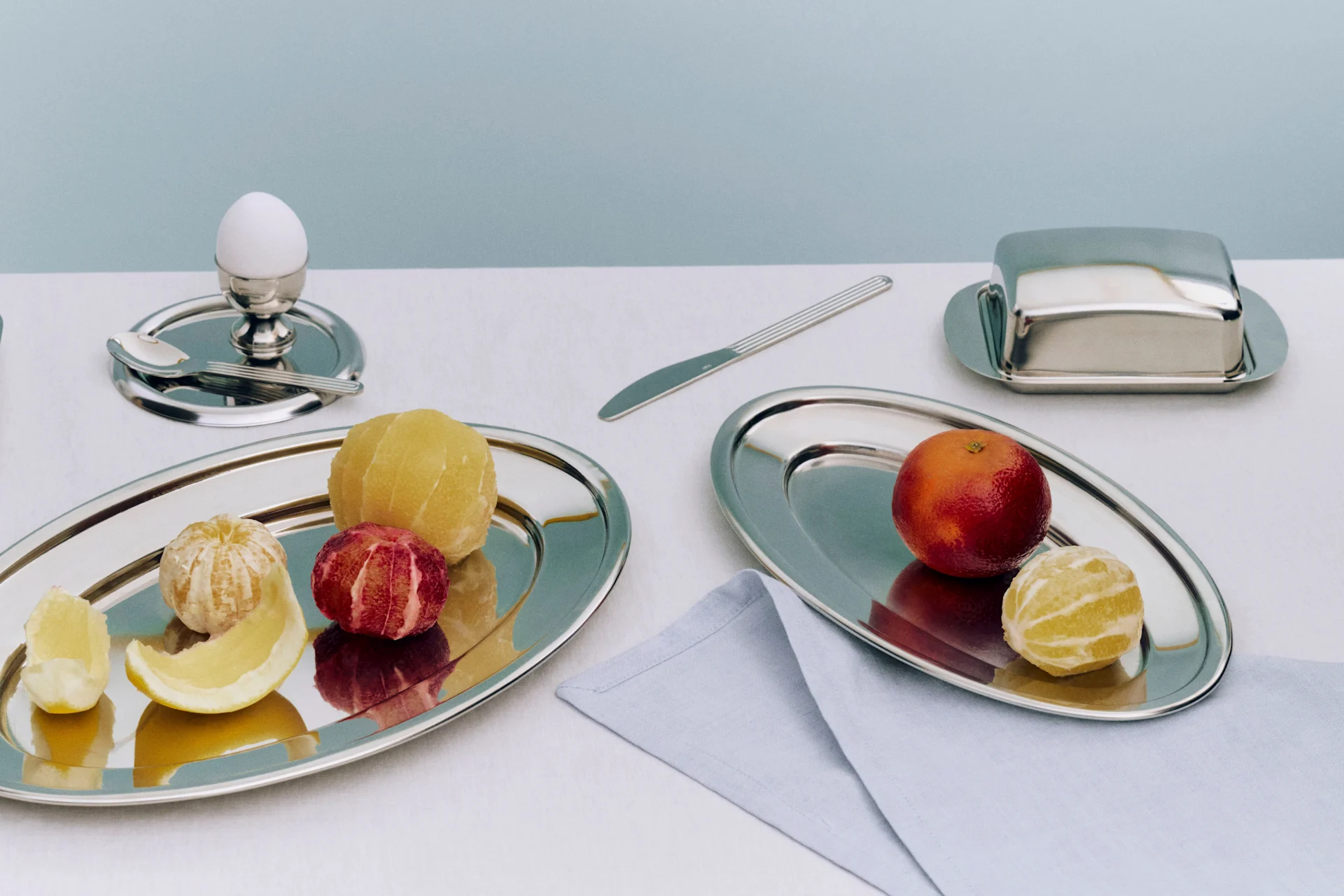 En frokostservering med to sølvfat med skrellede sitrusfrukter, et egg i en eggekopp, en smørskål og en kniv.