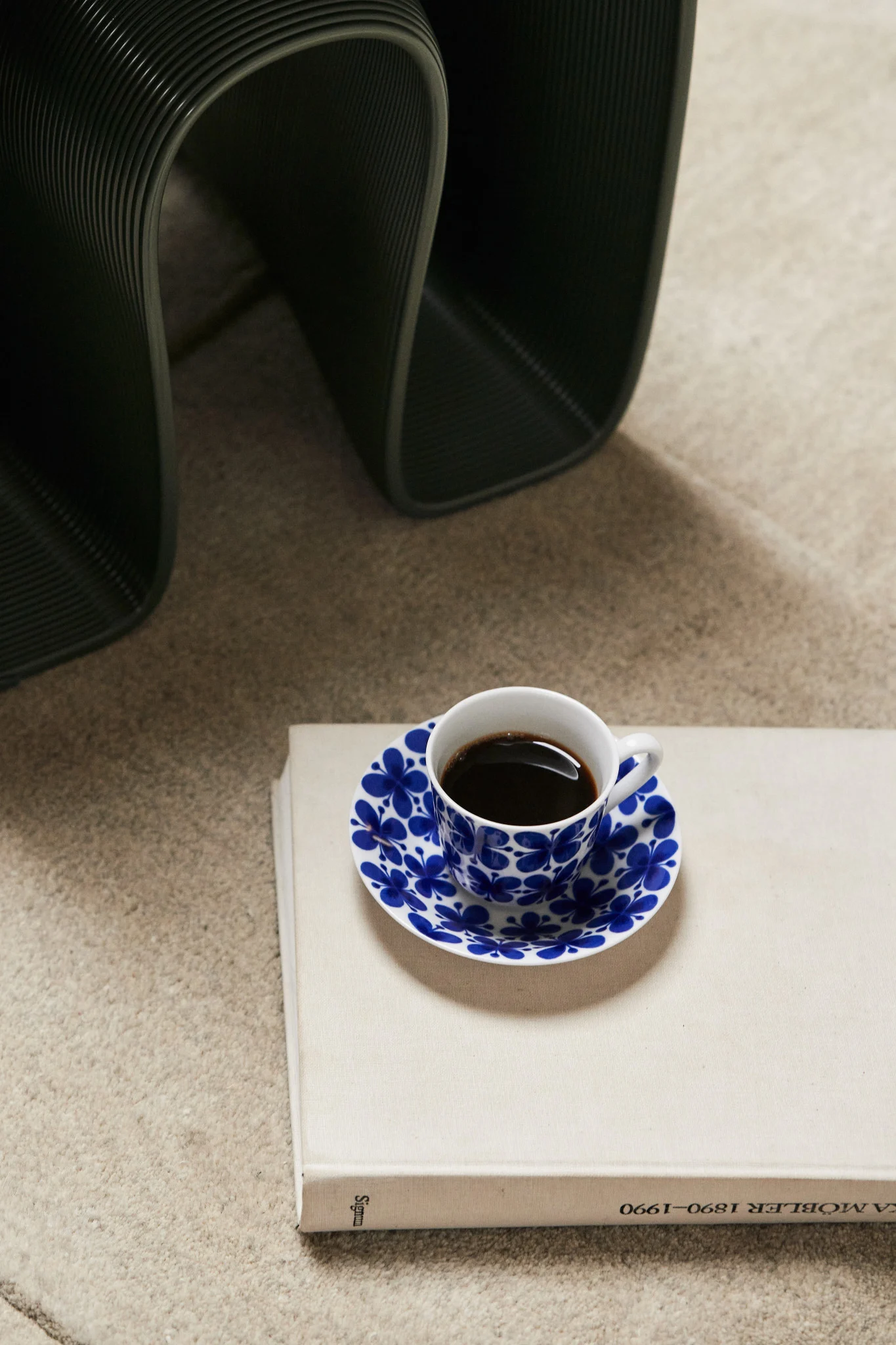 Kaffekopp med blått blomstret fat på en lys bok, mørkt ribbet objekt i bakgrunnen.