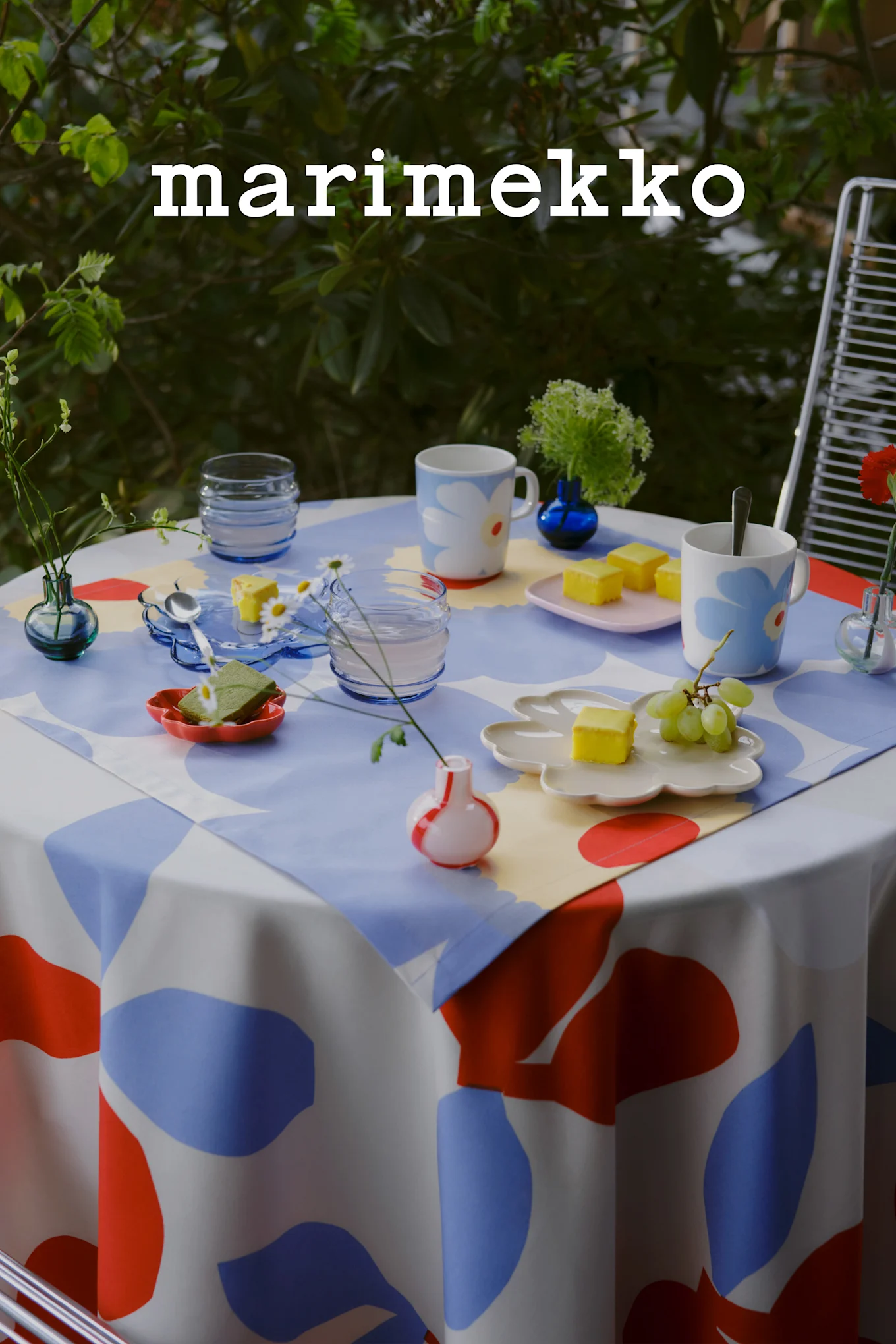 Marimekko-logo. Et utebord dekket med en blå, rød og hvit abstrakt duk, blomstrete krus, gule kaker, druer og små vaser.