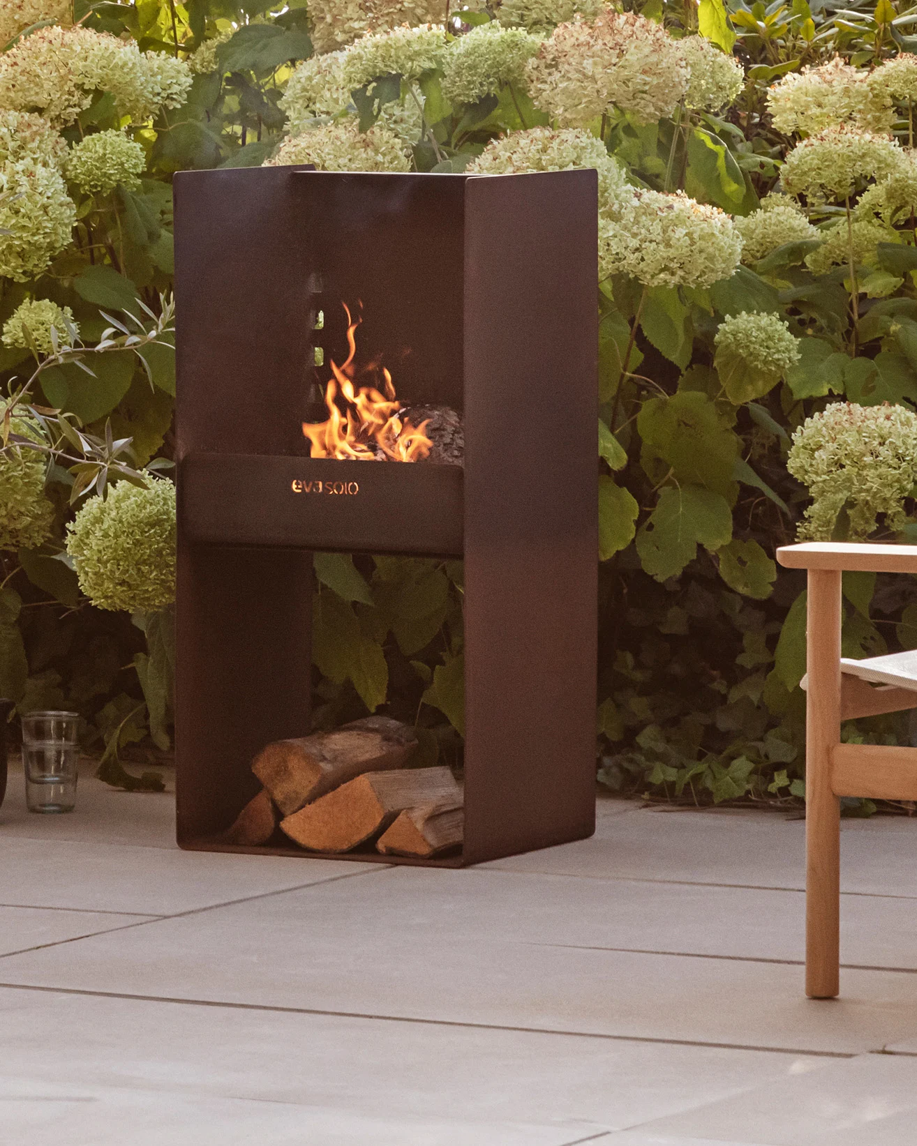 Moderne mørk metall utendørs peis med brennende ild, stablede vedkubber under, omgitt av grønne planter og en uteplass.