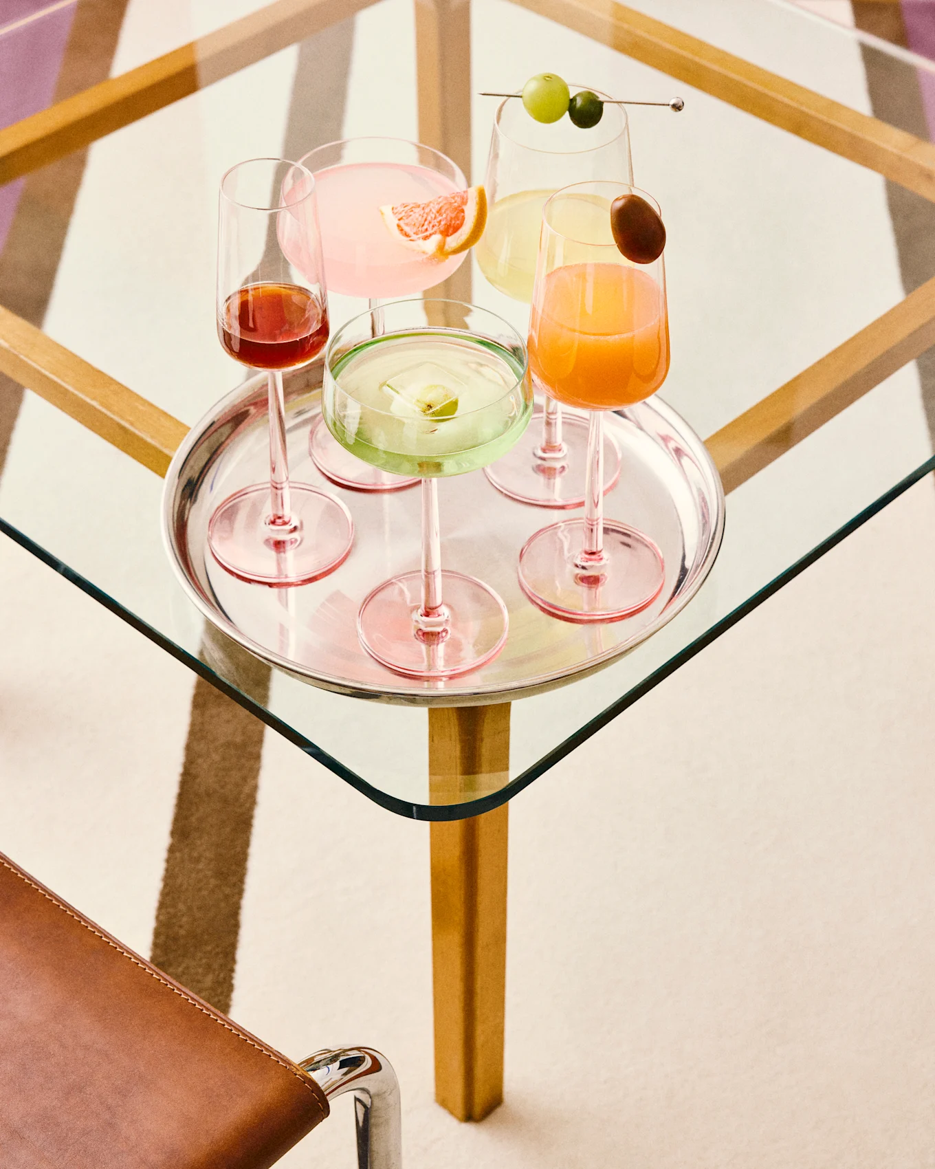 Fem fargerike cocktailer på et sølvbrett: rosa med grapefrukt, grønn med fruktis, gul med druer, oransje med garnityr og mørkerød, alle på et glassbord.