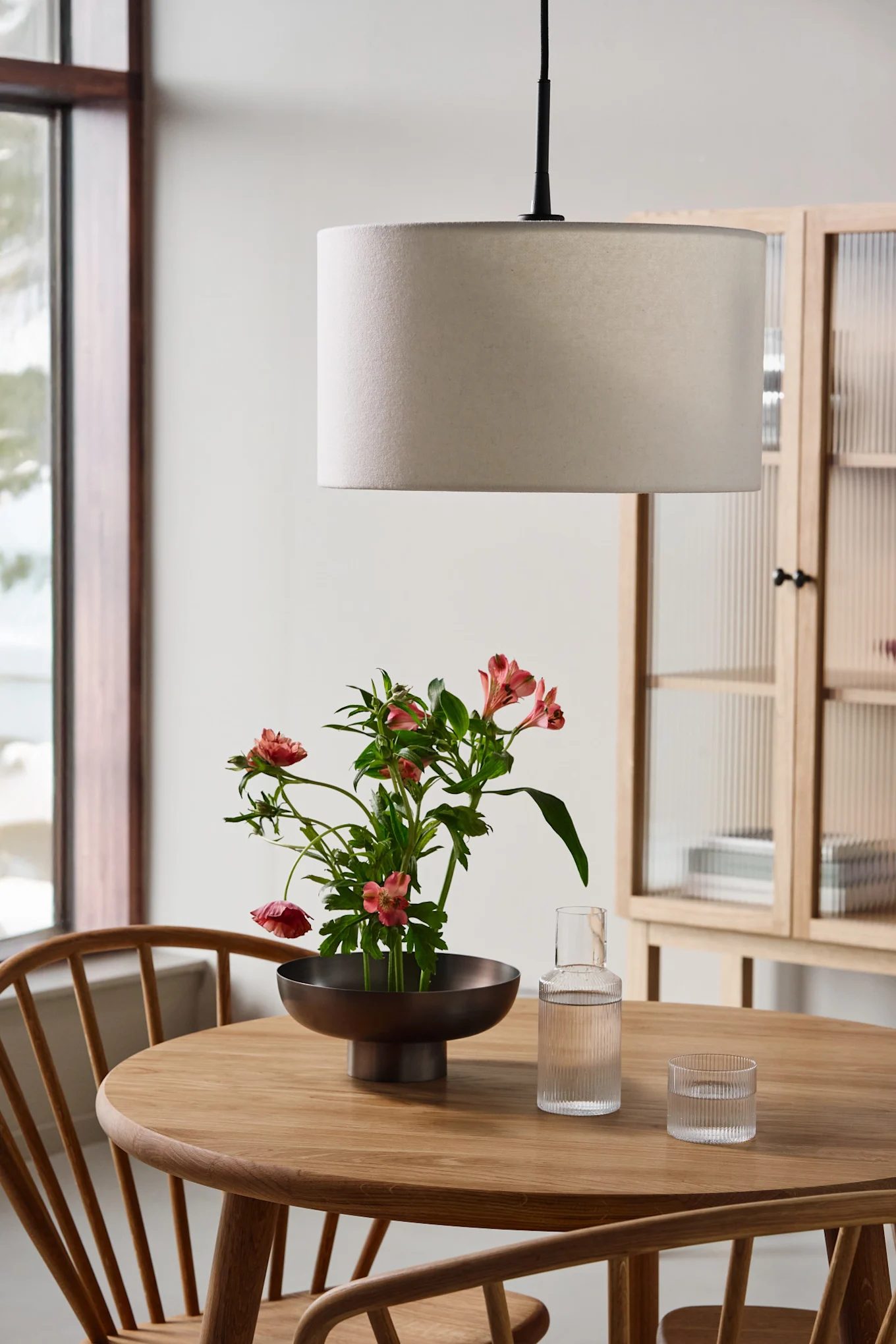 Et rundt trebord med en skål rosa blomster, en glasskaraffel, et glass og en trommependellampe.