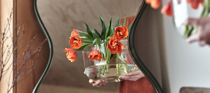 Et par hender bærer en vase med blomster arrangert i en klar glassvase reflektert i et buet speil fra Ekbacken Studios.
