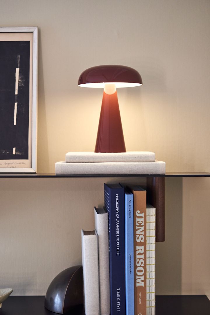 Como bærbar og oppladbar lampe i rødt fra &tradition på en stilren bokhylle omgitt av bøker.