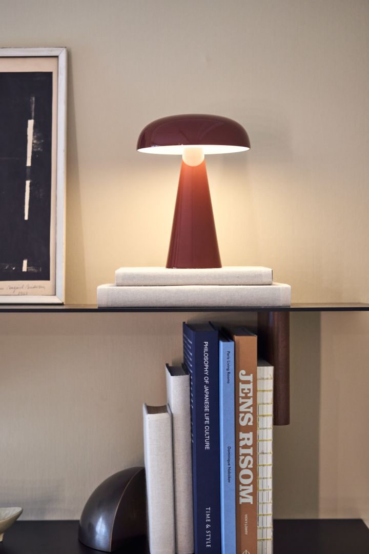 Como bærbar og oppladbar lampe i rødt fra &tradition på en stilren bokhylle omgitt av bøker.