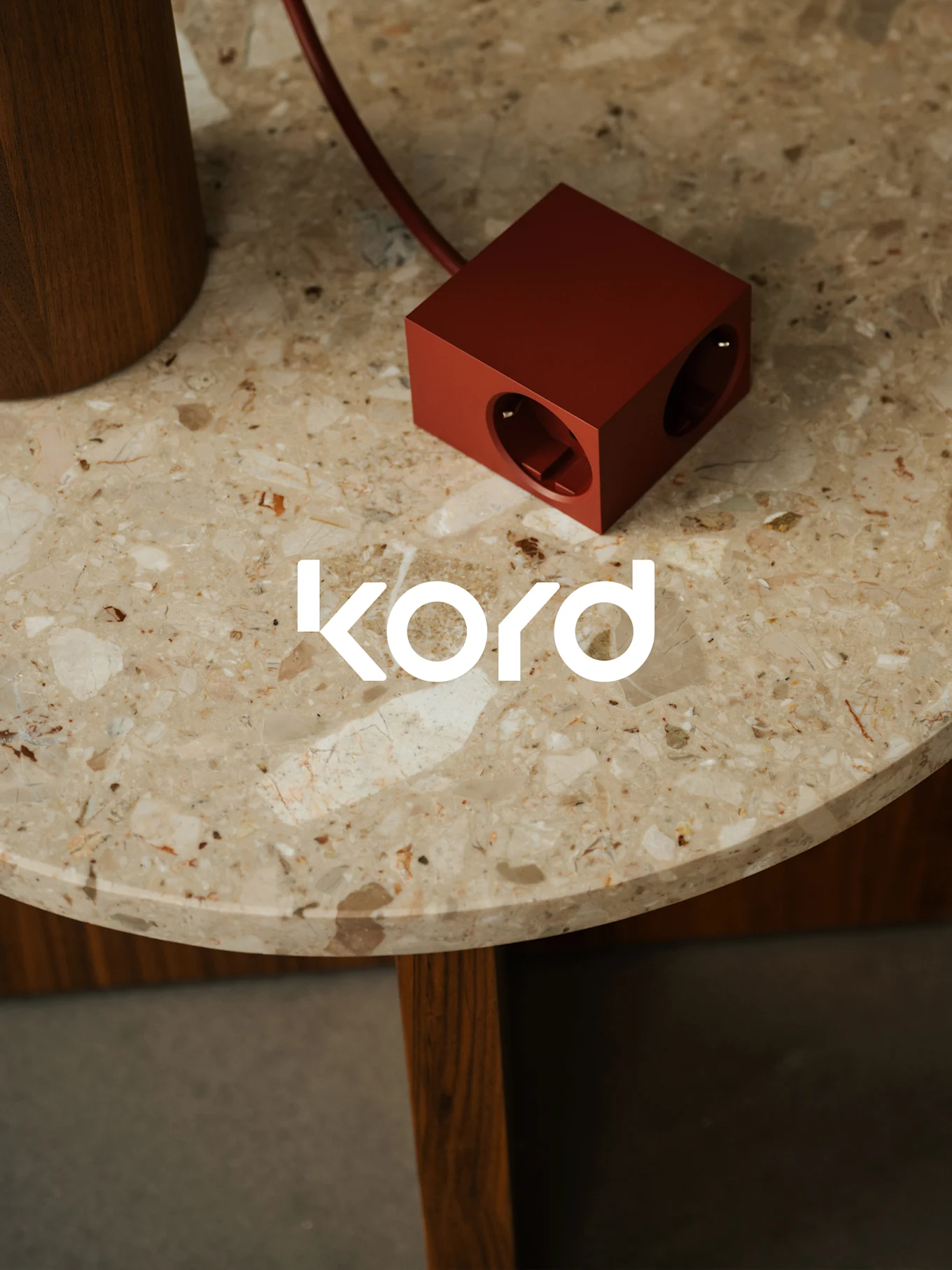 En mørkerød, kubeformet strømskinne med to synlige uttak hviler på et rundt marmorbord, med den hvite "kord"-logoen.