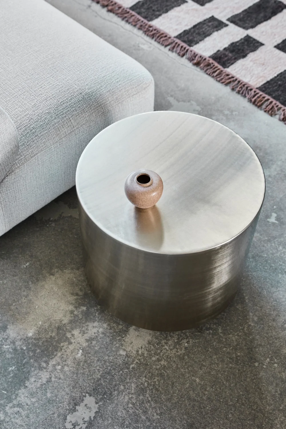 Rundt metallisk sidebord med en liten keramisk vase, ved siden av en lys grå sofa og mønstret teppe på betonggulv.