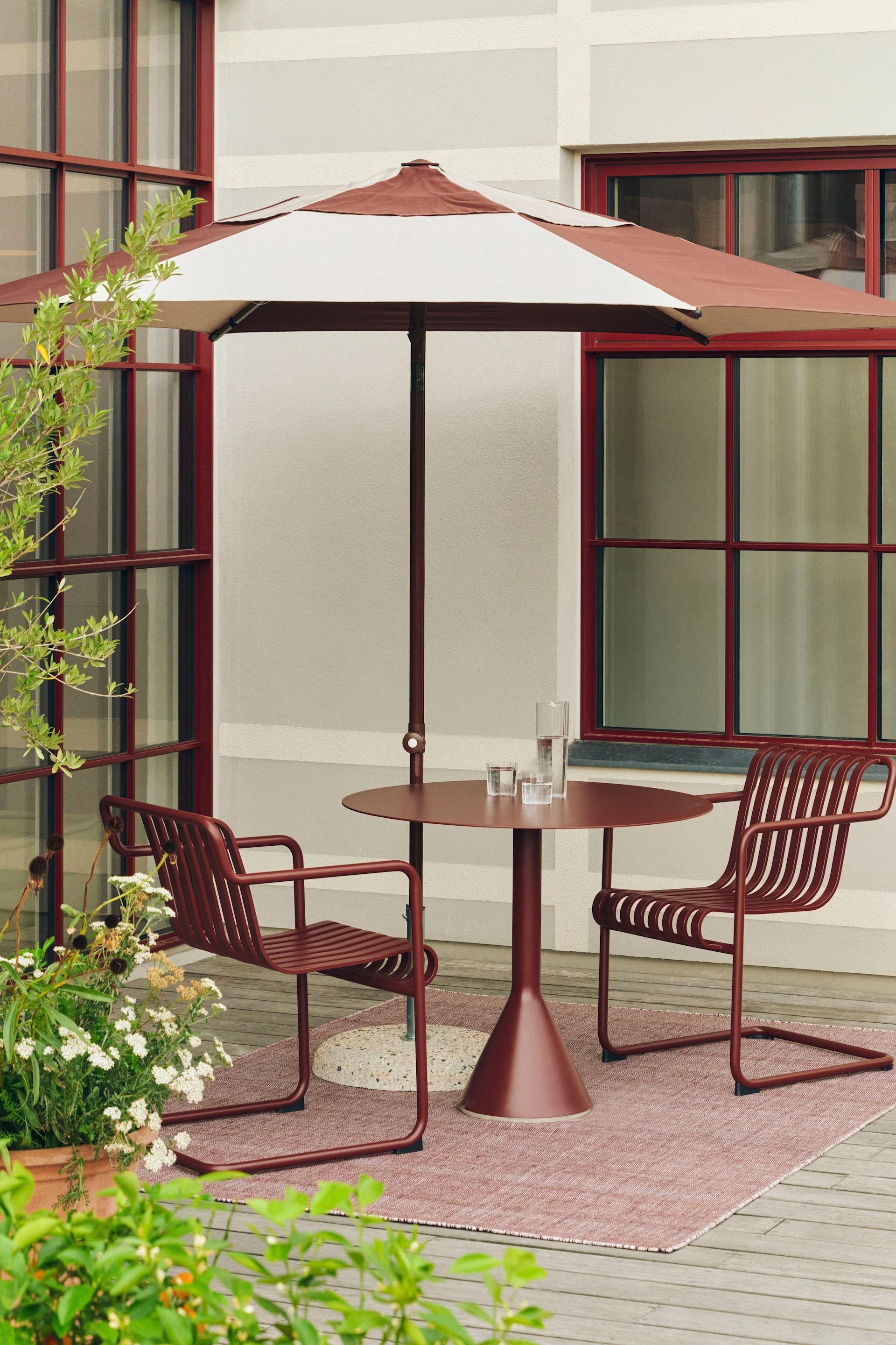 Uteplass med rødbrun stripete parasoll, matchende rundt bord og to lamellstoler på et rosa teppe.