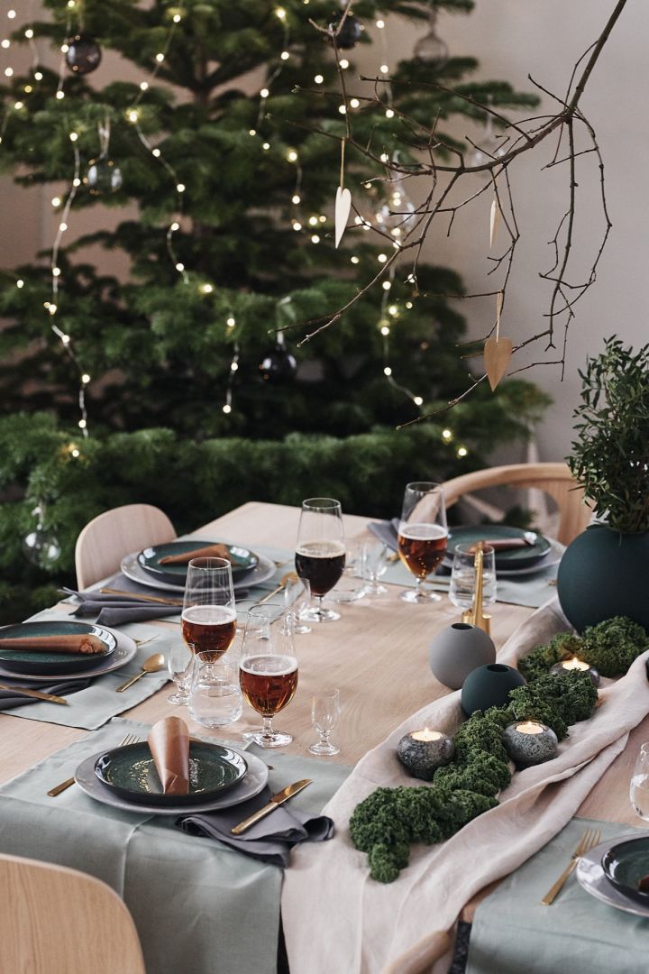 Det er dekket til julemiddag med en grønn borddekking med grønne tallerkener, gullbestikk og grønne vaser.