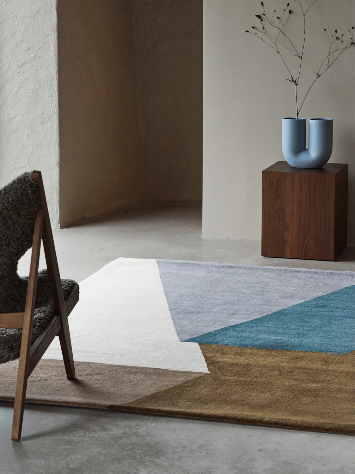 Moderne rom med et geometrisk teppe i hvitt, grått, turkis og brunt, en U-formet blå vase på en trepiedestall og en lodden stol.