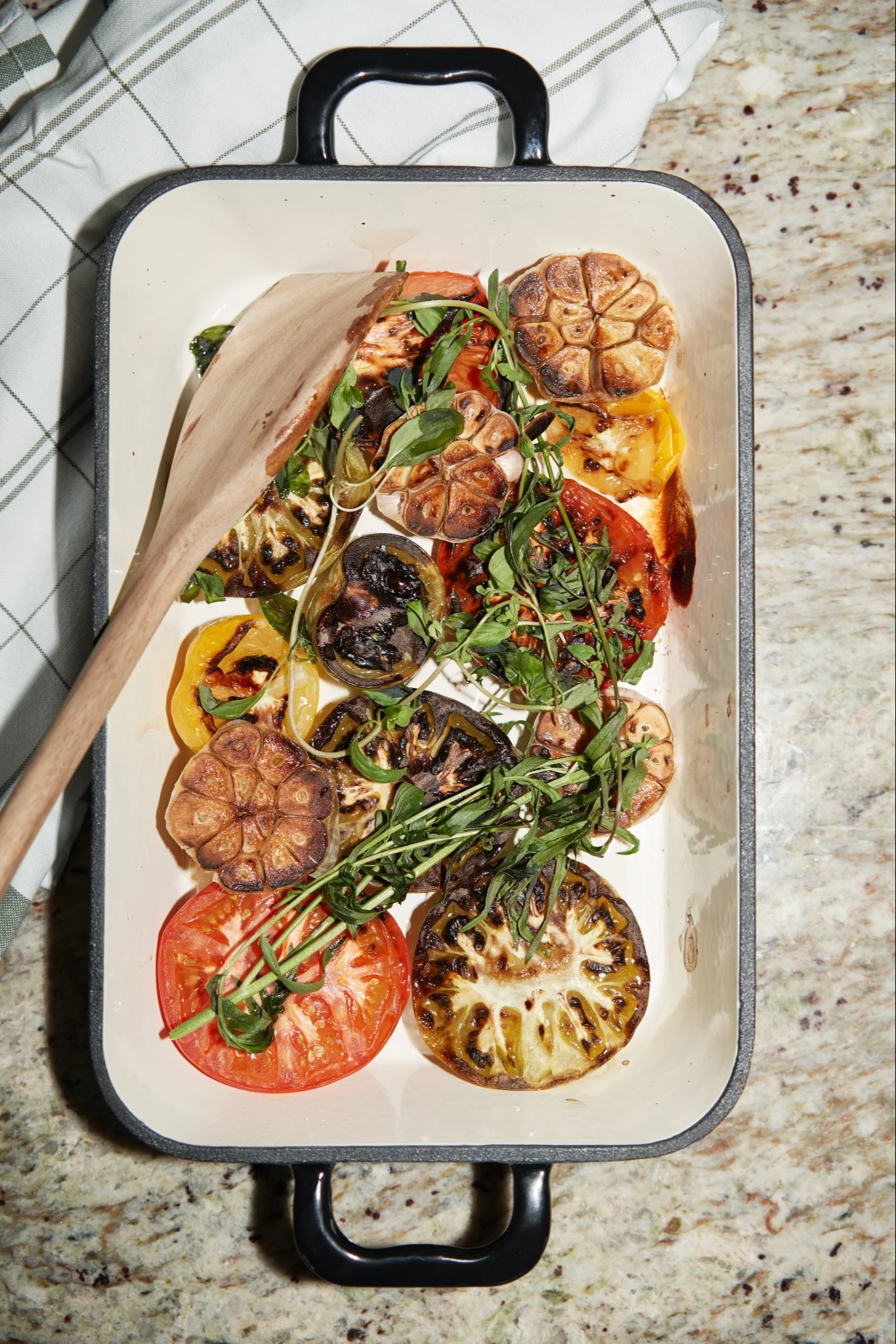 En hvit bakeform fylt med grillede tomater, aubergine, stekt hvitløk og friske grønne urter, med en tresleiv.