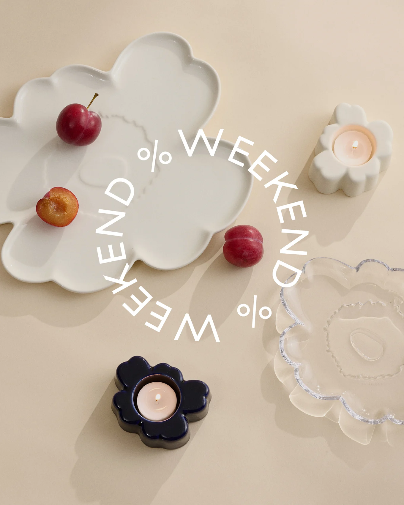Dekorative tallerkener og lysestaker i hvitt, blått og klart glass med plommer på en beige bakgrunn. Hvit tekst sier 'Weekend %'.