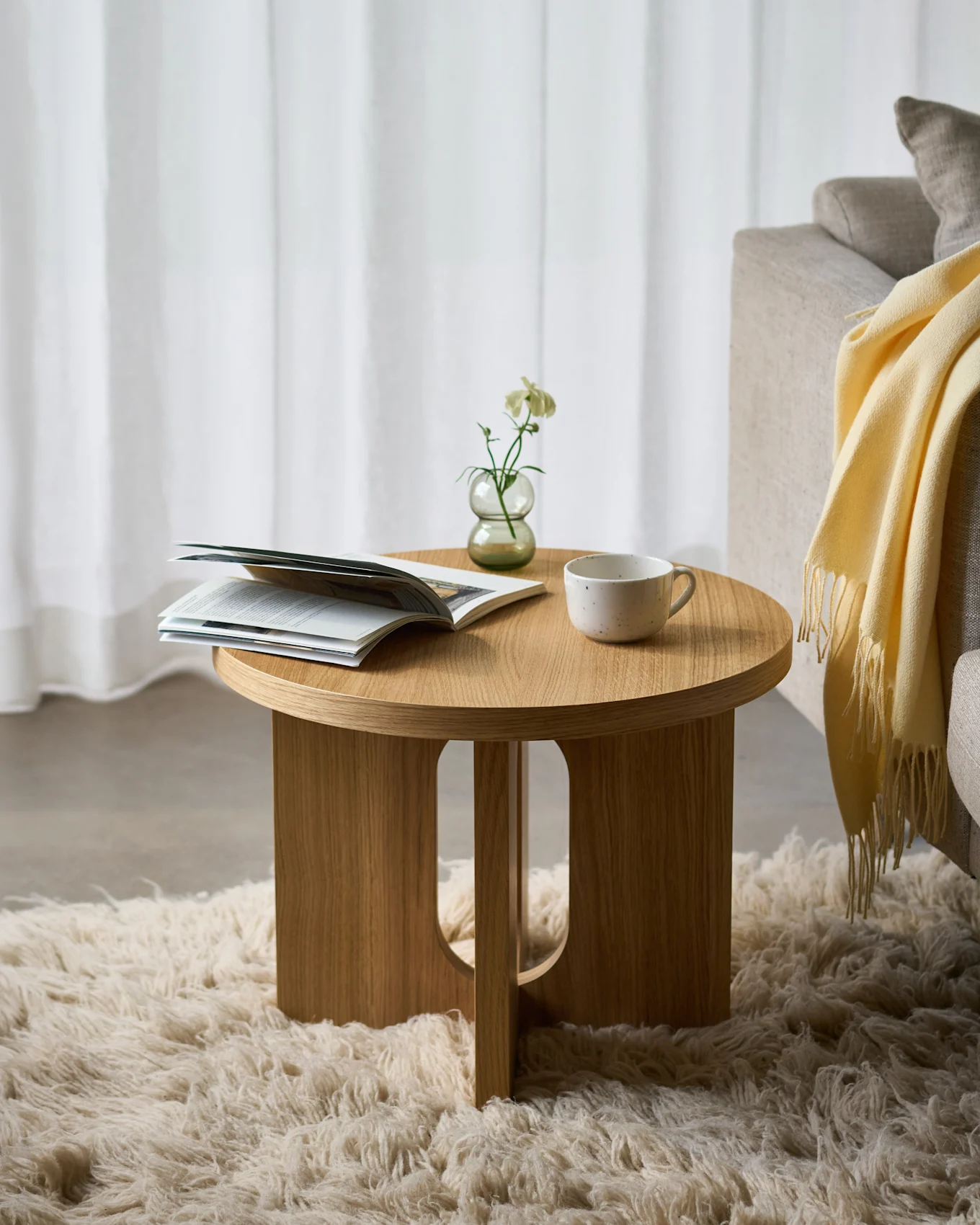 Rundt sidebord i tre på et mykt teppe, med et åpent magasin, en hvit blomst i en vase og en keramikkop, ved siden av en sofa.
