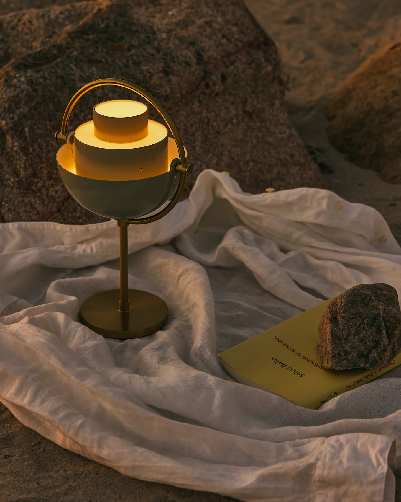 En moderne hvit og messing bordlampe med glødende lys, en gul bok og en stein på et hvitt tøy på en steinete overflate.
