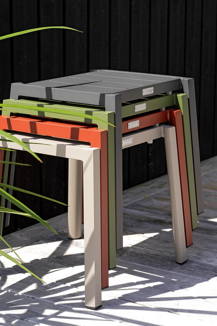 En stabel med fire rektangulære utendørs sidebord i mørkegrått, olivengrønt, terrakottaoransje og lys beige, på et tredekke med et mørkt gjerde.