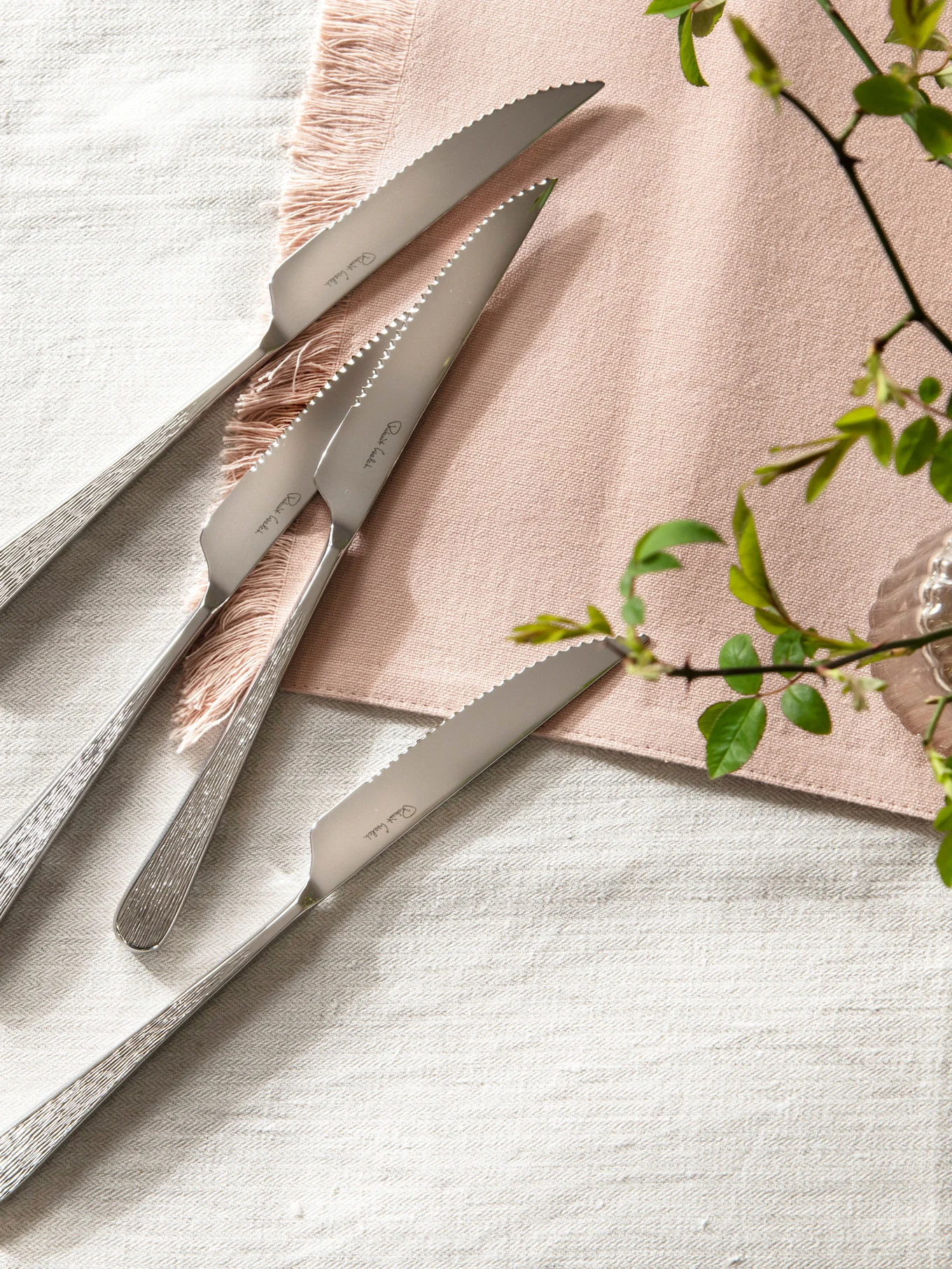 Fire biffkniver i rustfritt stål med taggete blader og teksturerte håndtak på en linserviet med en rosa serviett.