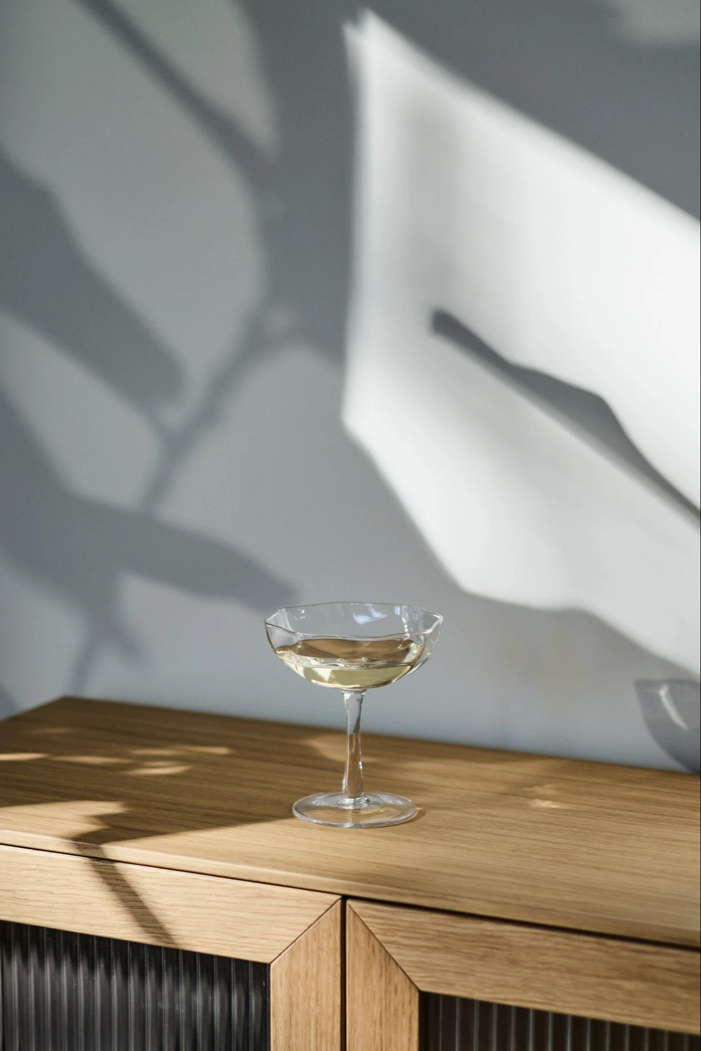 Et coupeglass med en lys drink på et treskap, med abstrakte skygger på veggen.