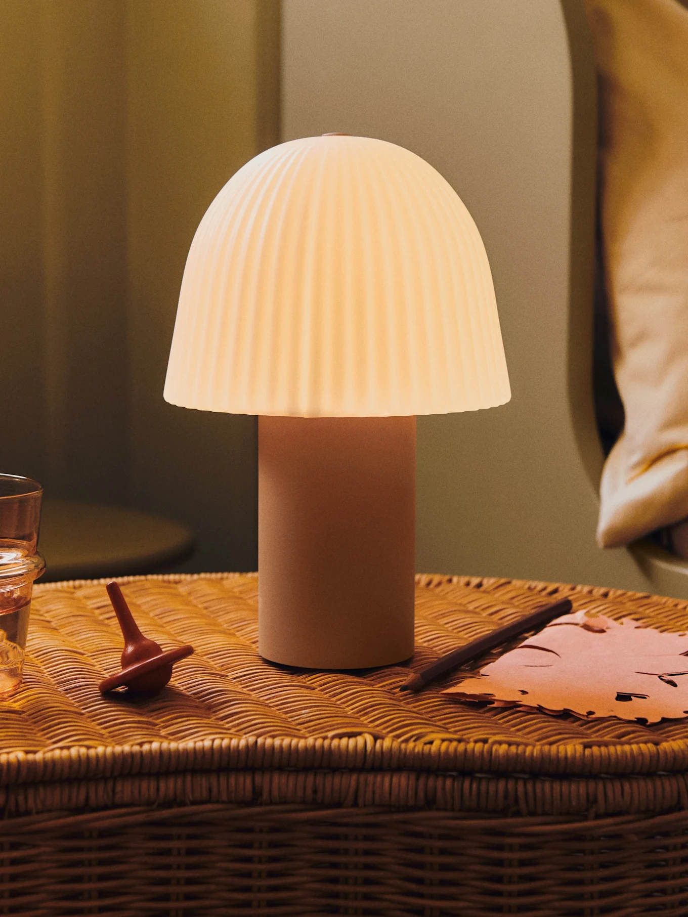 En glødende sopplampe med plissert skjerm på et flettet nattbord med et glass vann, en snurrebass og papir.