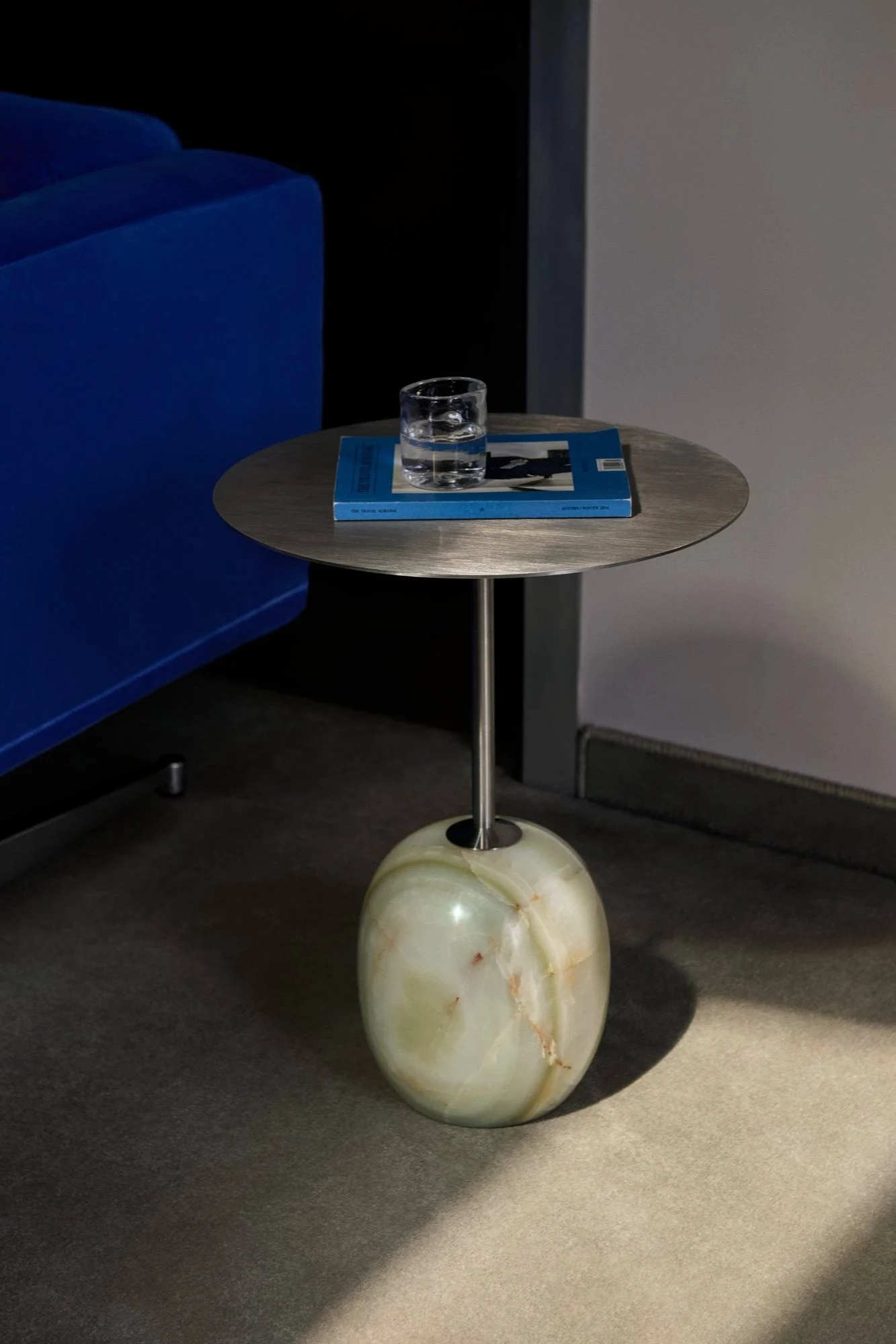 Sidebord med børstet metalltopp, avrundet grønn onyxbase, glass vann, blå bok, ved siden av en blå sofa.