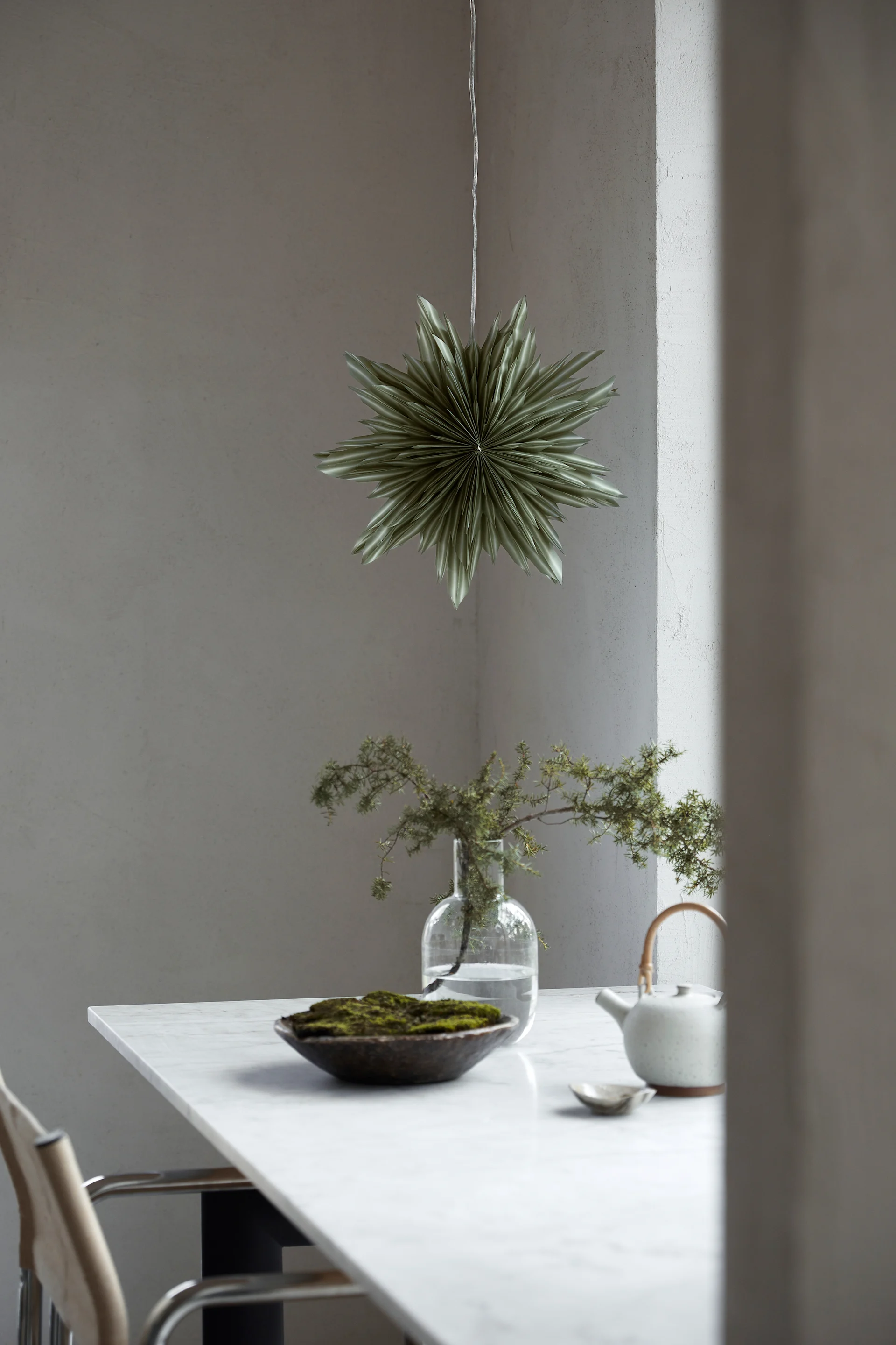 Helena, en grønn adventsstjerne i papir, henger i vinduet over en grønn potteplante.
