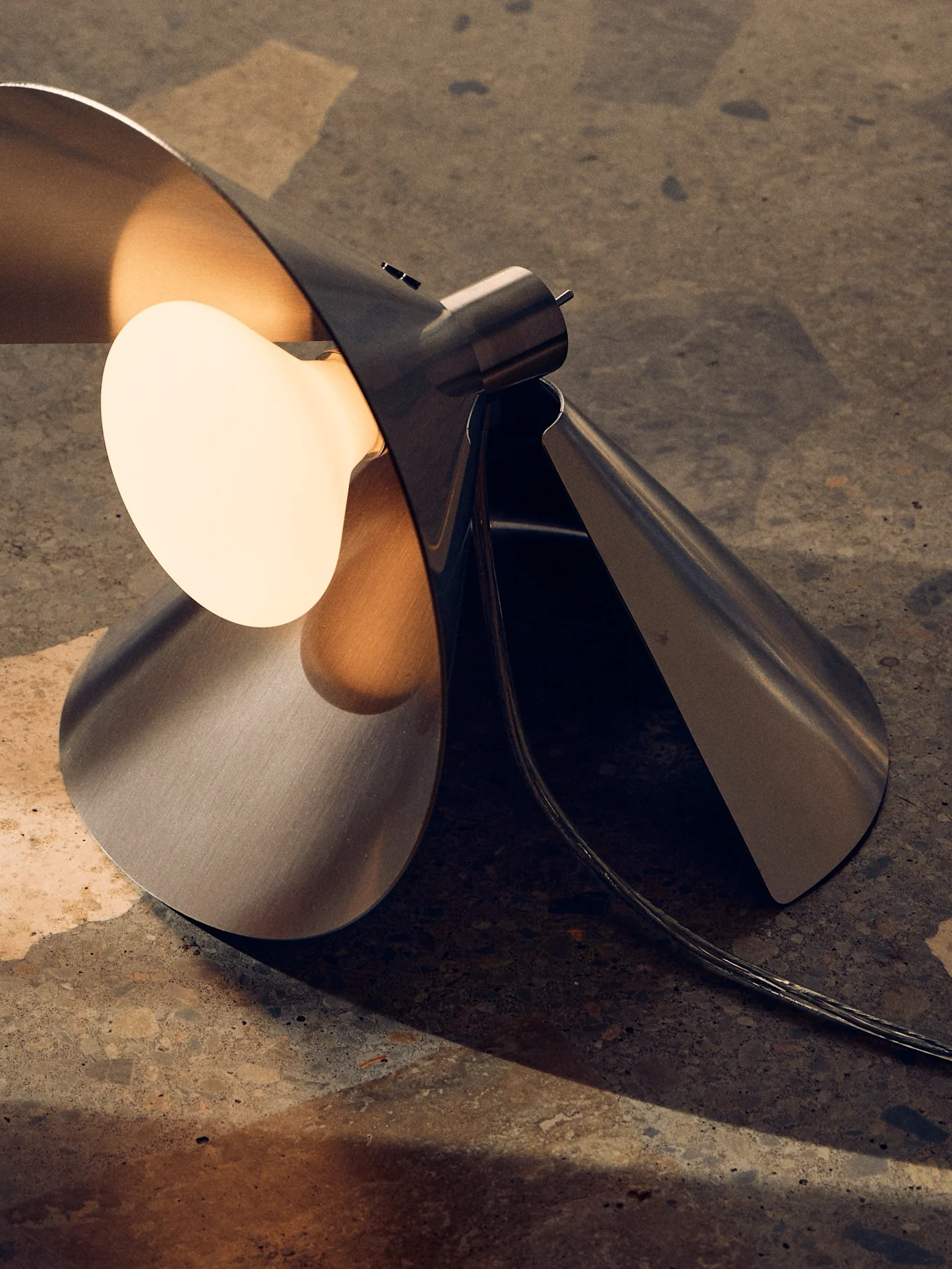 Moderne bordlampe i børstet metall med skulpturelt, foldet design og en varm, eksponert pære på en teksturert overflate.