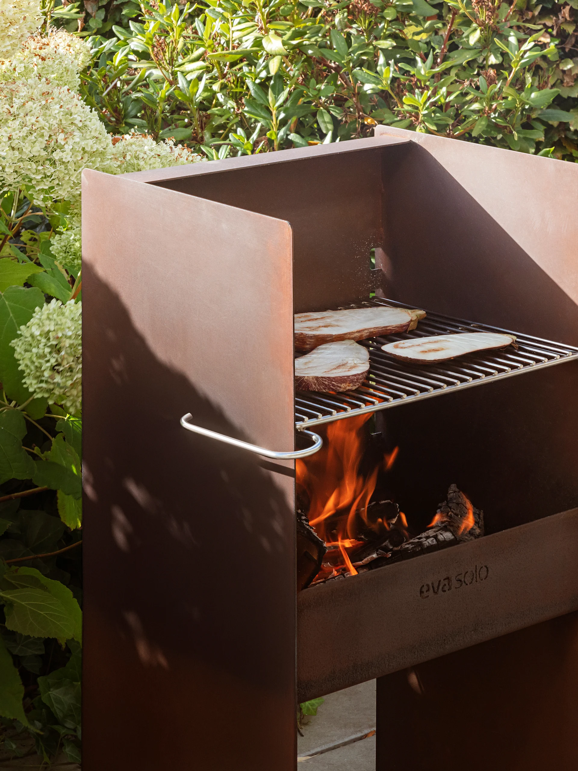 Rusten metall utendørsgrill fra Eva Solo med aubergine på risten, brennende ild og frodig løvverk.