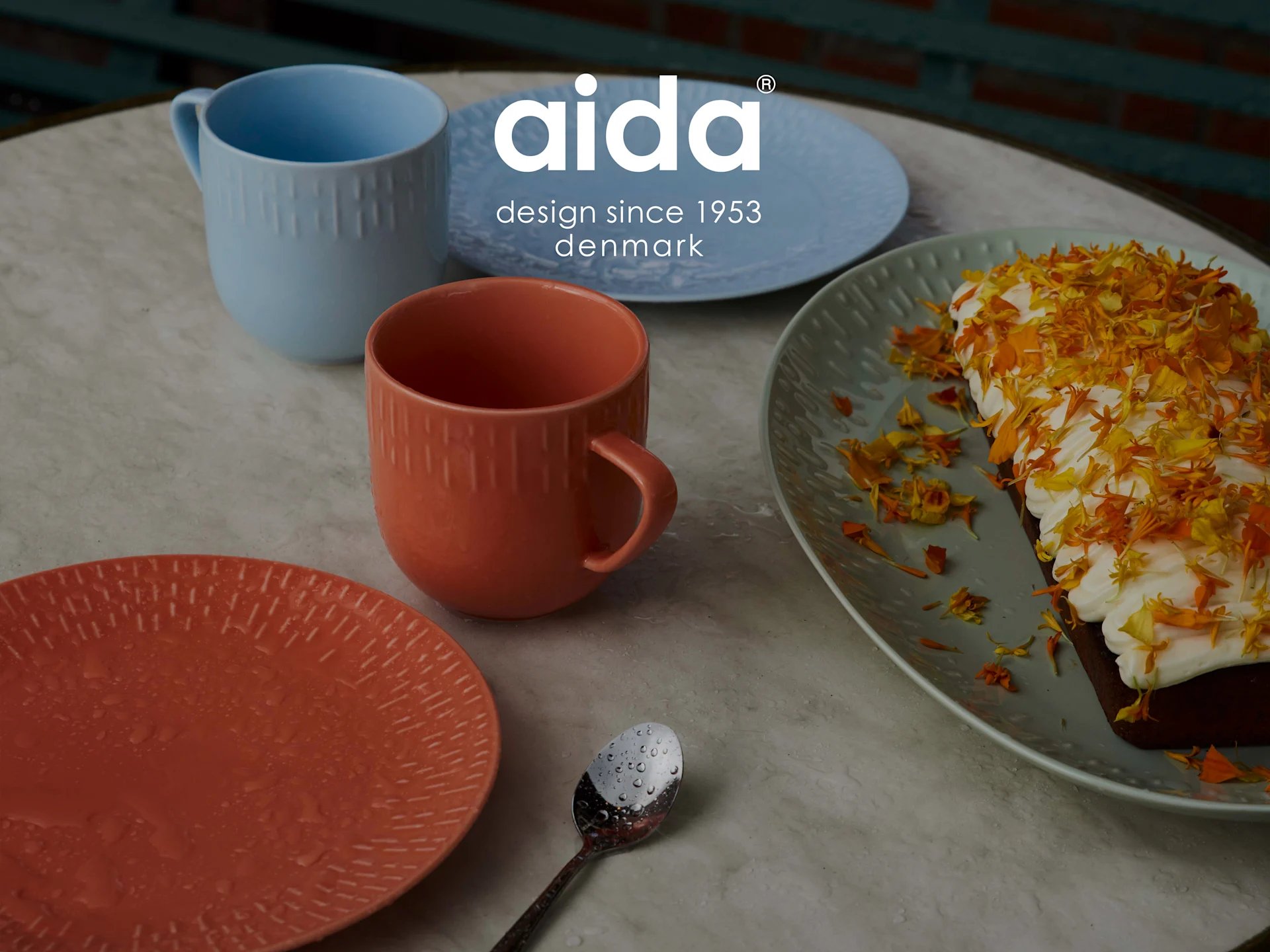 Aida krus og tallerkener i blått og oransje, med et kakestykke toppet med krem og gule kronblader, og en skje med vanndråper.
