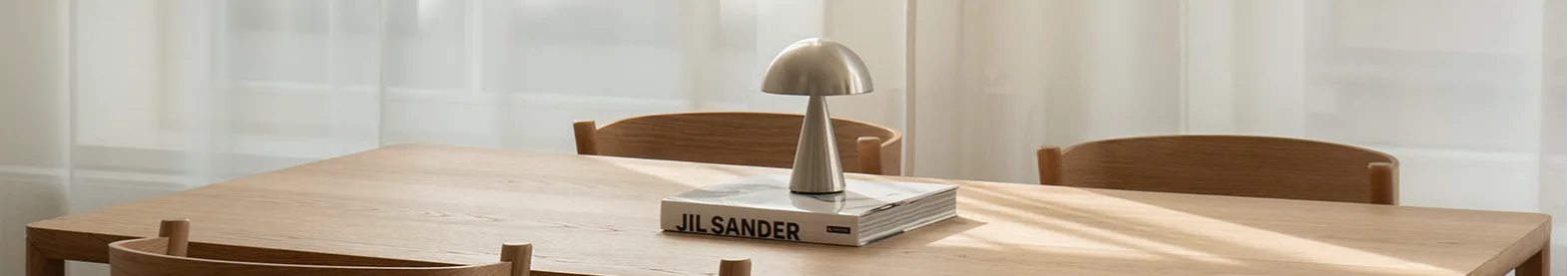 Spisebord i lyst tre med fire stoler. Sølvfarget sopplampe og bok på bordet. Sollys gjennom gjennomsiktige hvite gardiner.