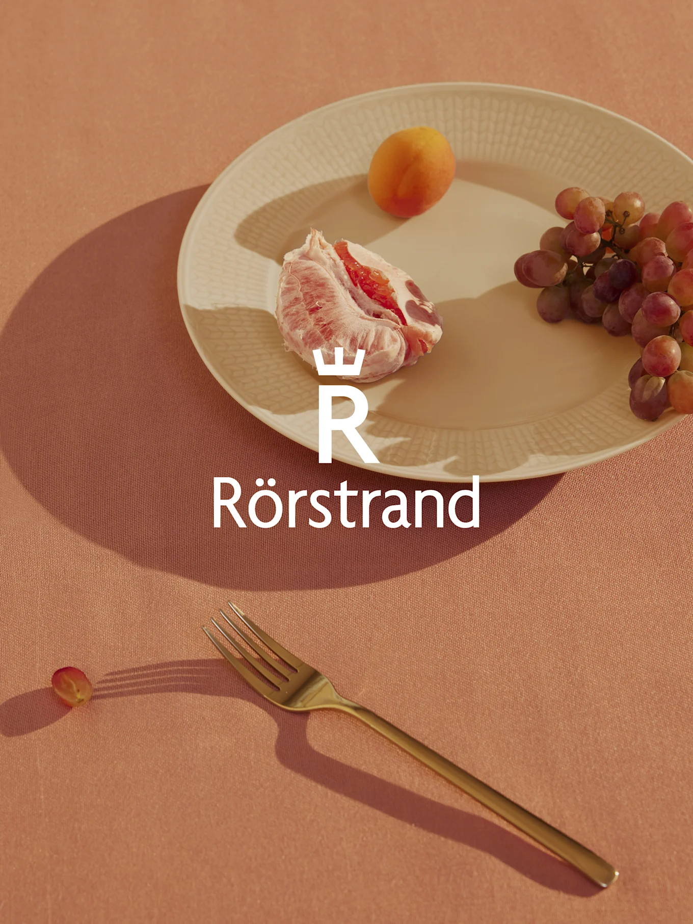 Rörstrand-logo over en tallerken med grapefrukt, abrikos og druer, en gaffel og en enkelt drue på en ferskenfarget duk.