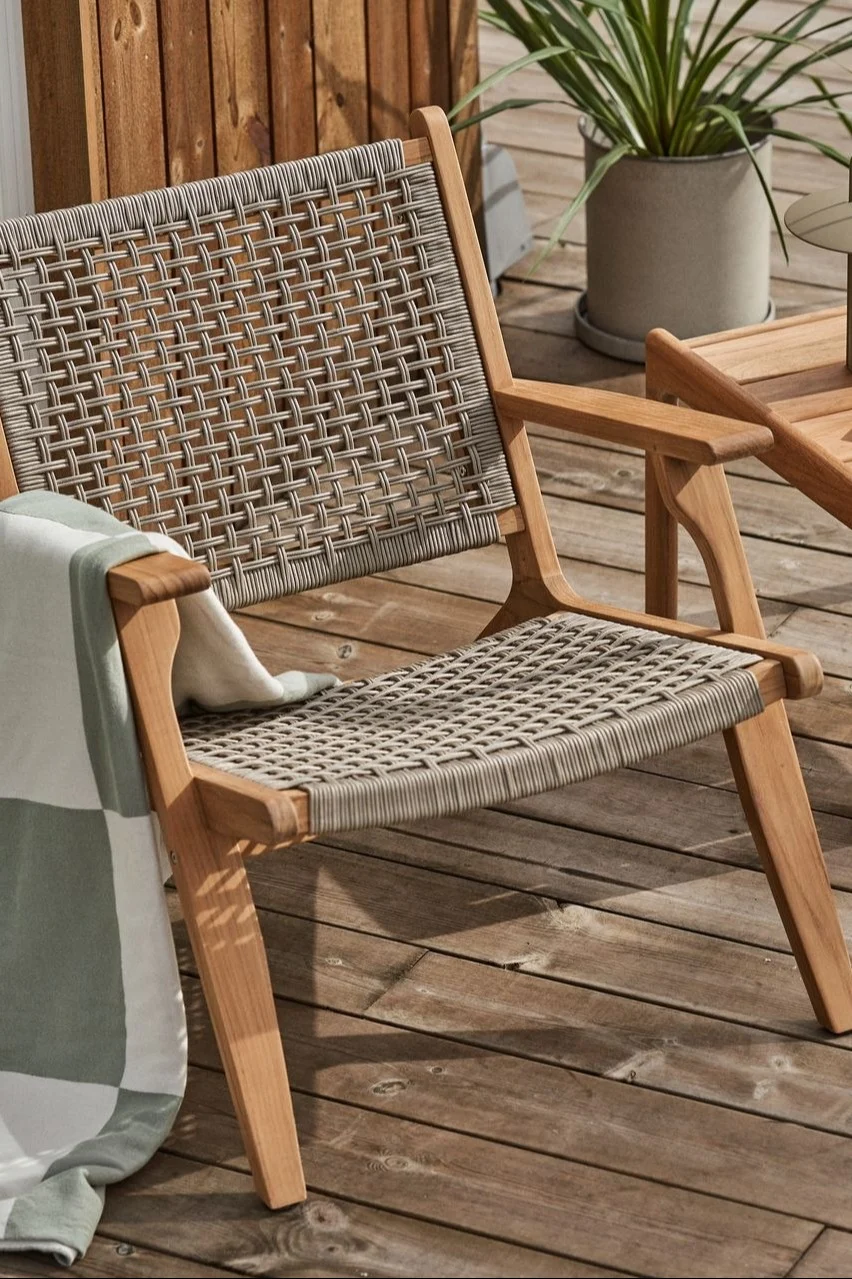 Utestol i tre med flettede grå stropper og et stripete teppe på en treterrasse.