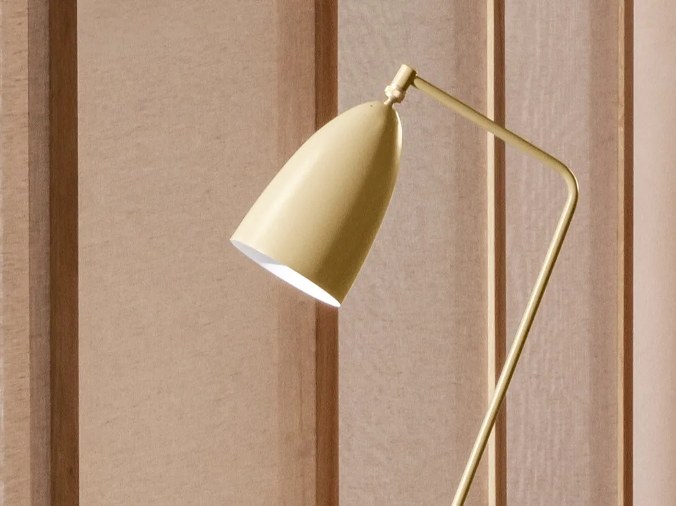 Et nærbilde av en gulaktig-gull moderne lampe med et konisk hode vinklet nedover, som sender ut lys mot en lysbrun panelvegg.