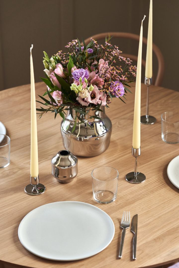 En speilblank vase i rustfritt stål står midt på et bord med en bukett lilla blomster.