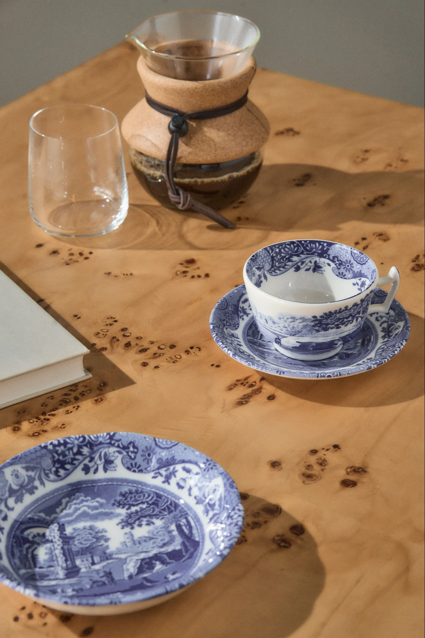 En pour-over kaffetrakter, et tomt glass, en blå- og hvitmønstret tekopp på et fat, og en matchende tallerken på et lyst trebord.