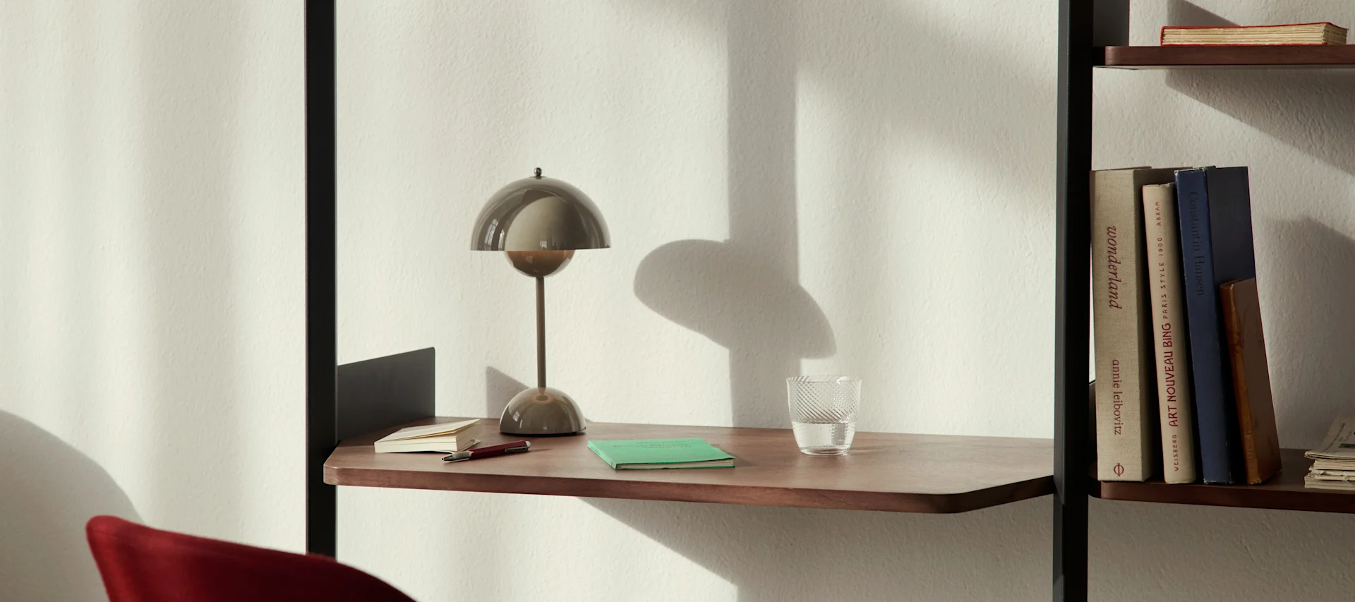 En moderne soppformet bordlampe, et glass vann og notatbøker på en trehylle ved siden av en bokhylle.