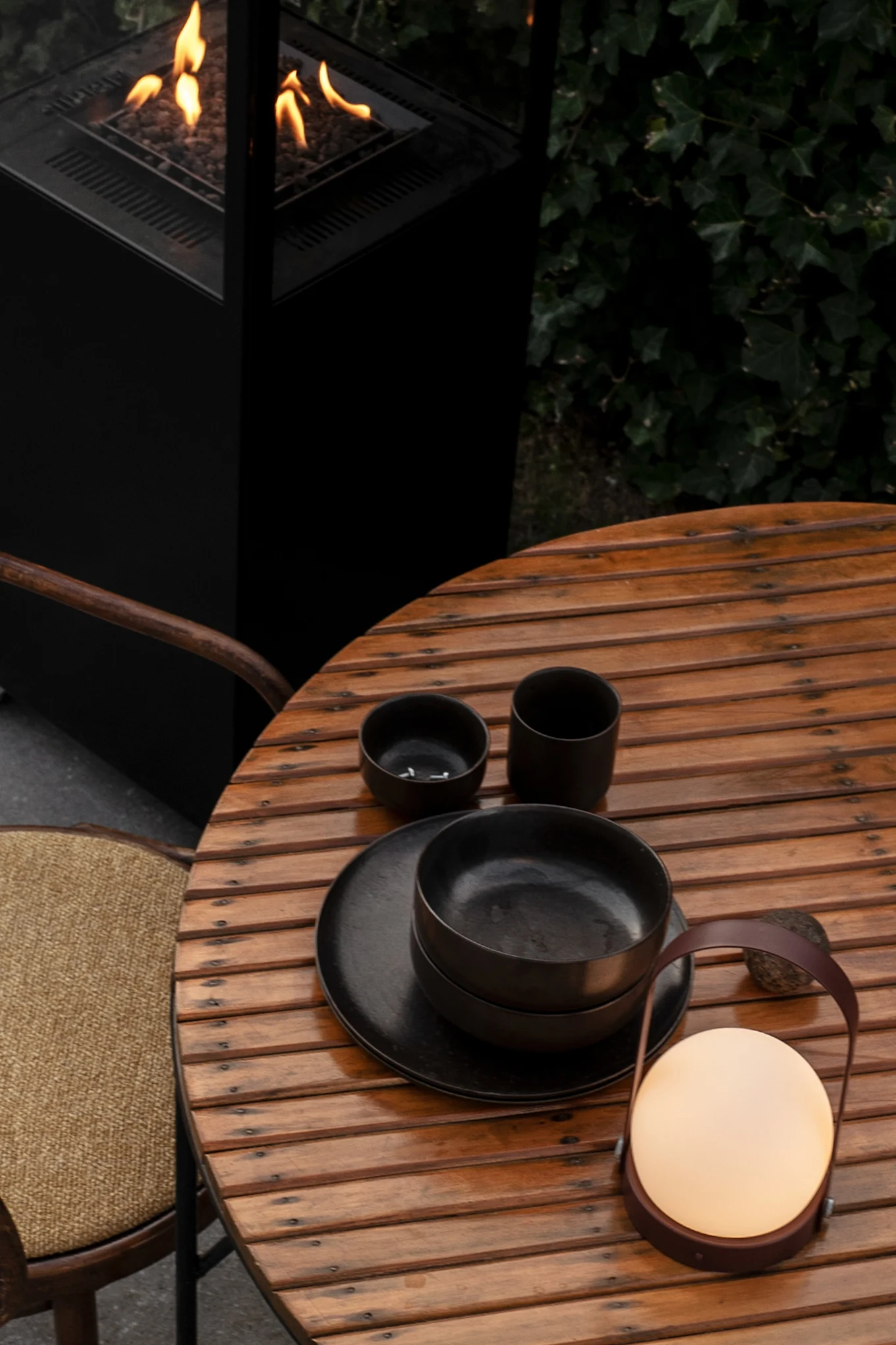 Carrie-lampe i Burned Red på et hagebord i tre, ved siden av et svart servise – med stol og bålpanne i bakgrunnen.