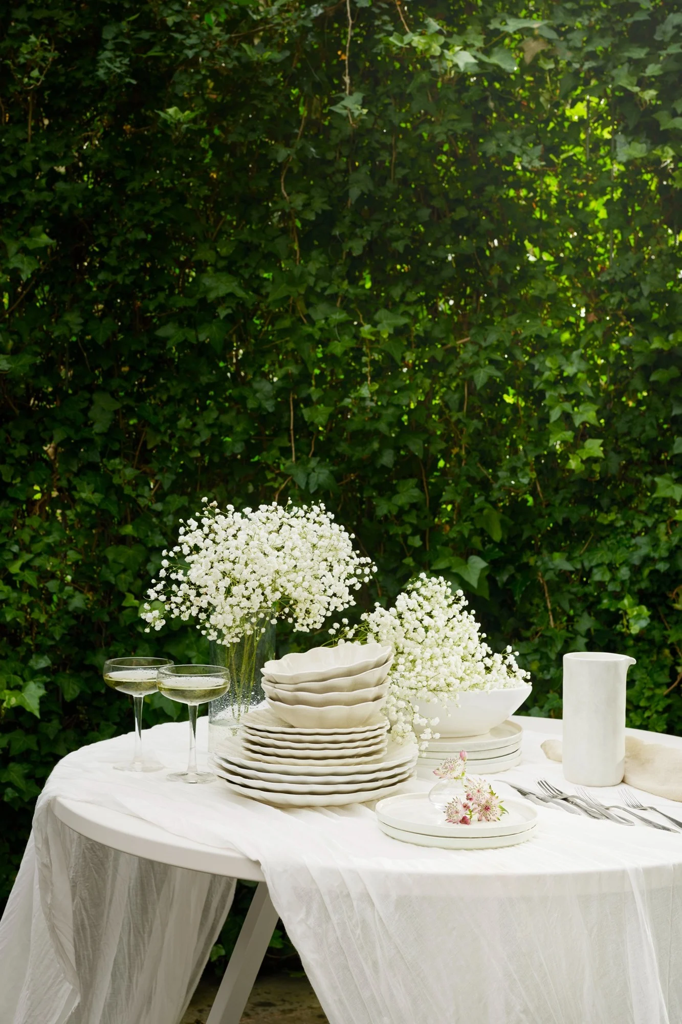 Utendørs bord dekket med hvitt servise, brudeslørblomster, champagneglass og bestikk på en hvit duk, mot frodige grønne blader.