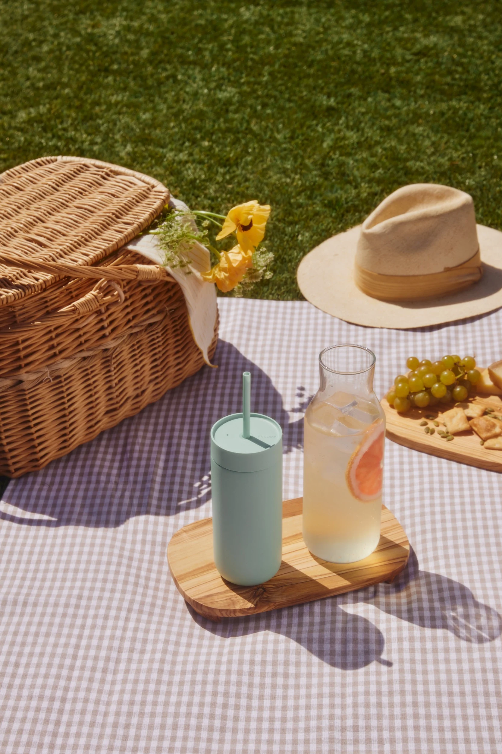 Piknik på et rutete teppe: flettet kurv, lysegrønn kopp, grapefruktdrikk, druer og kjeks på gresset.