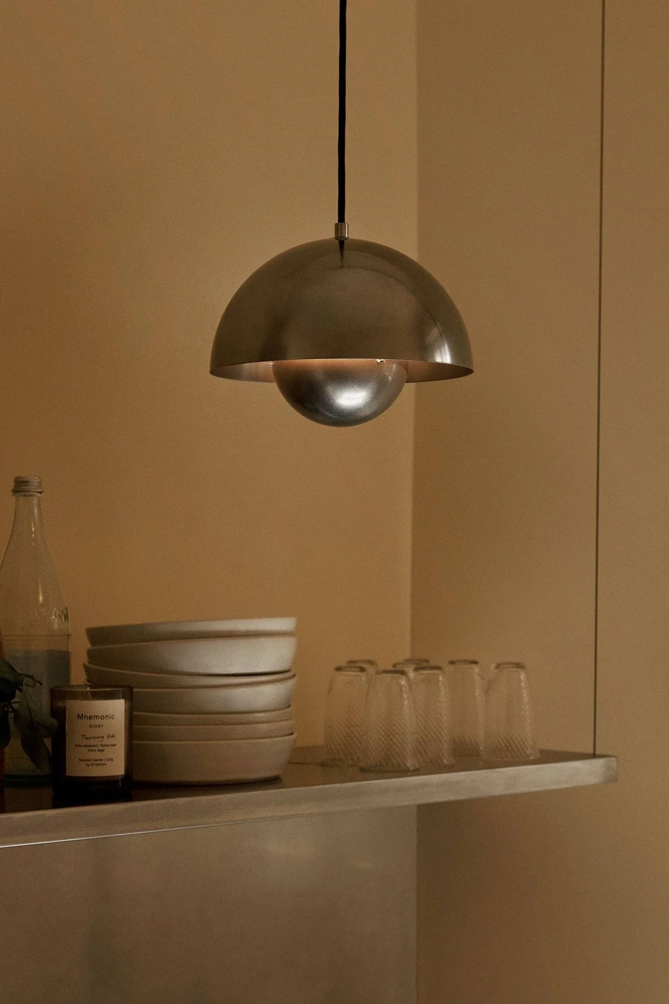 Metallisk kuppelpendellampe lyser opp hylle med stablede skåler, glass, flaske og lys i et varmtonet rom.