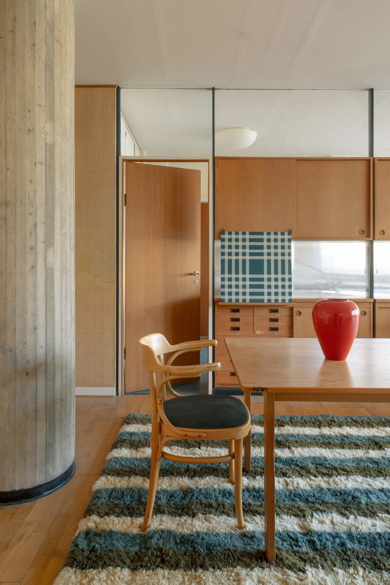 Moderne interiør med spisebord i tre, stol, rød vase og et stripete teppe. En betongsøyle rammer inn treskap og en åpen dør.