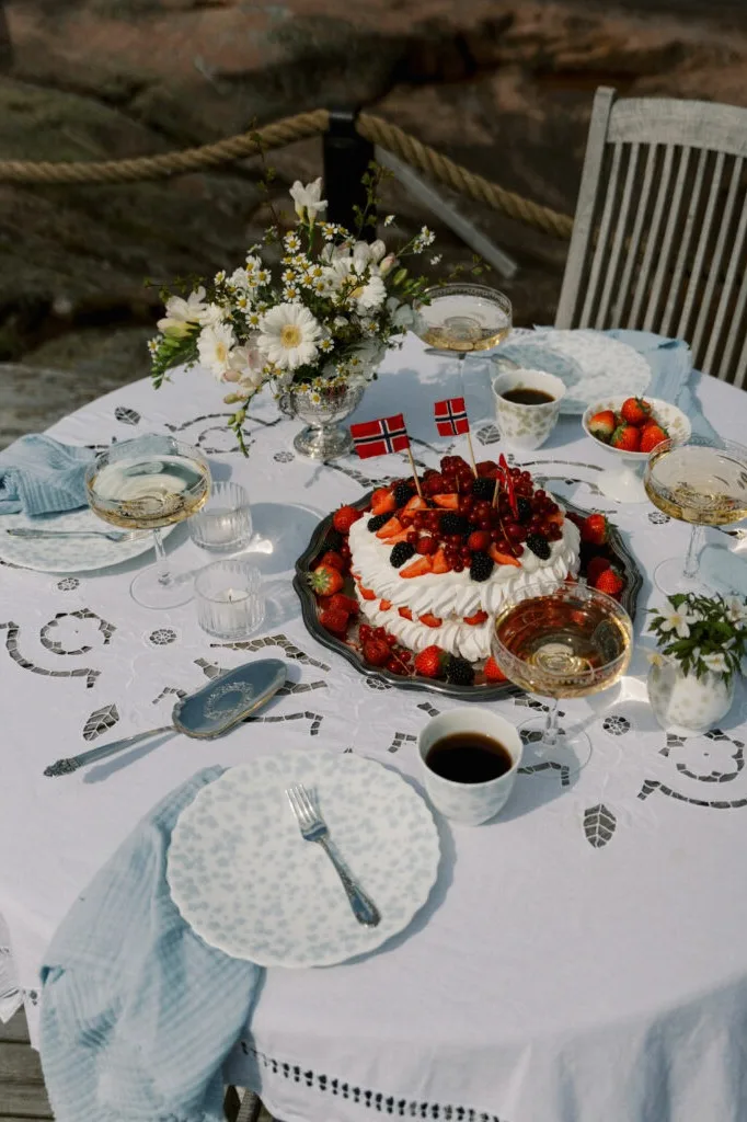 Utendørs bord med hvit brodert duk, pavlova kake med bær og norske flagg, champagne og hvite blomster.